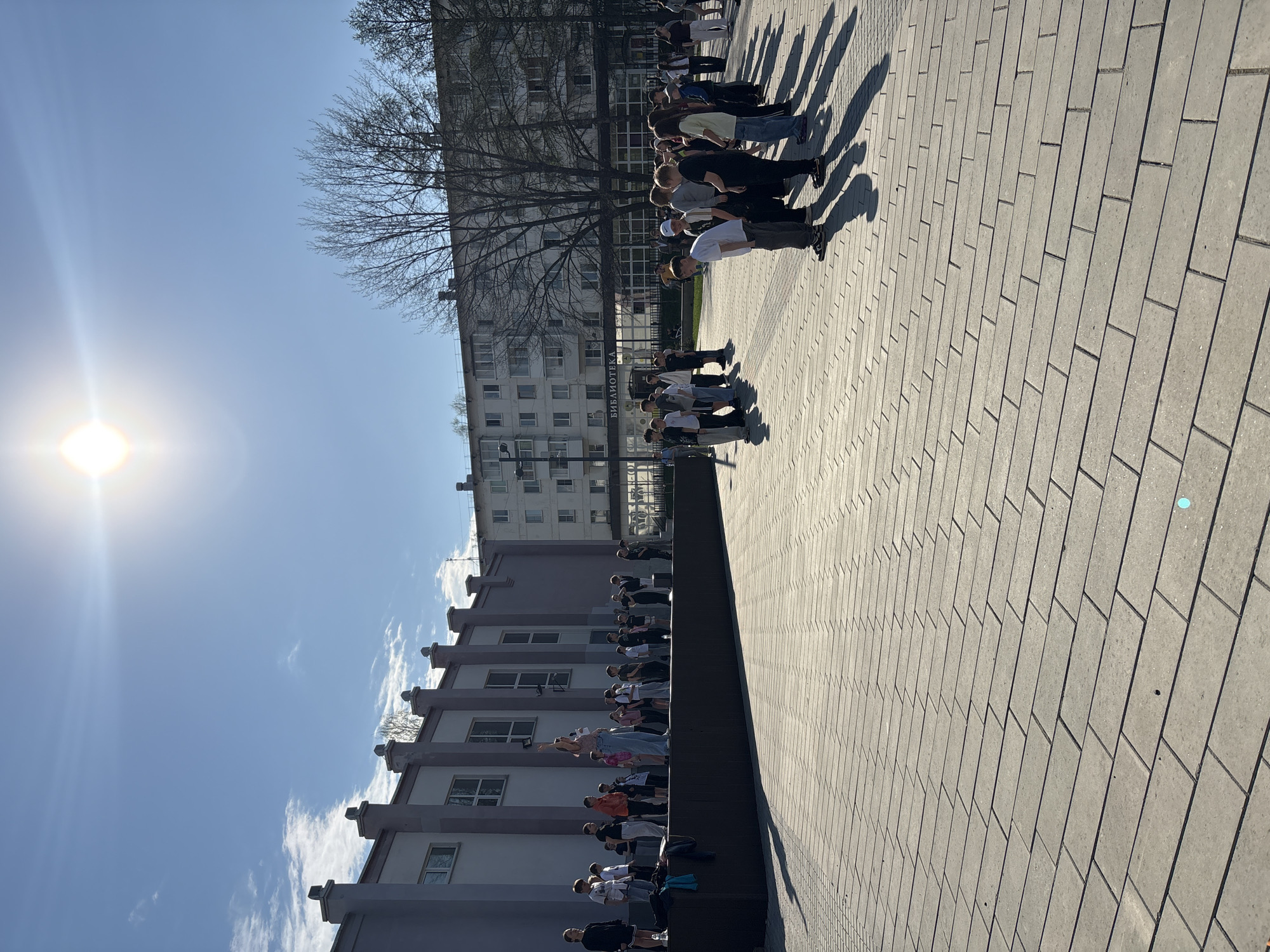 The image depicts a group of people standing or leaning over a low wall or ledge in an urban setting. They appear to be positioned in a line, possibly observing or participating in some activity. The scene is situated in Chaikovsky, Russia as there is a sign that reads "Библиотека" which translates to "Library" in Russian.
The setting includes buildings, possibly apartments, a paved area with a distinctive pattern, and a clear blue sky with a visible sun. The weather is sunny, casting long shadows of the people onto the pavement. The individuals in the group vary in clothing and appearance.