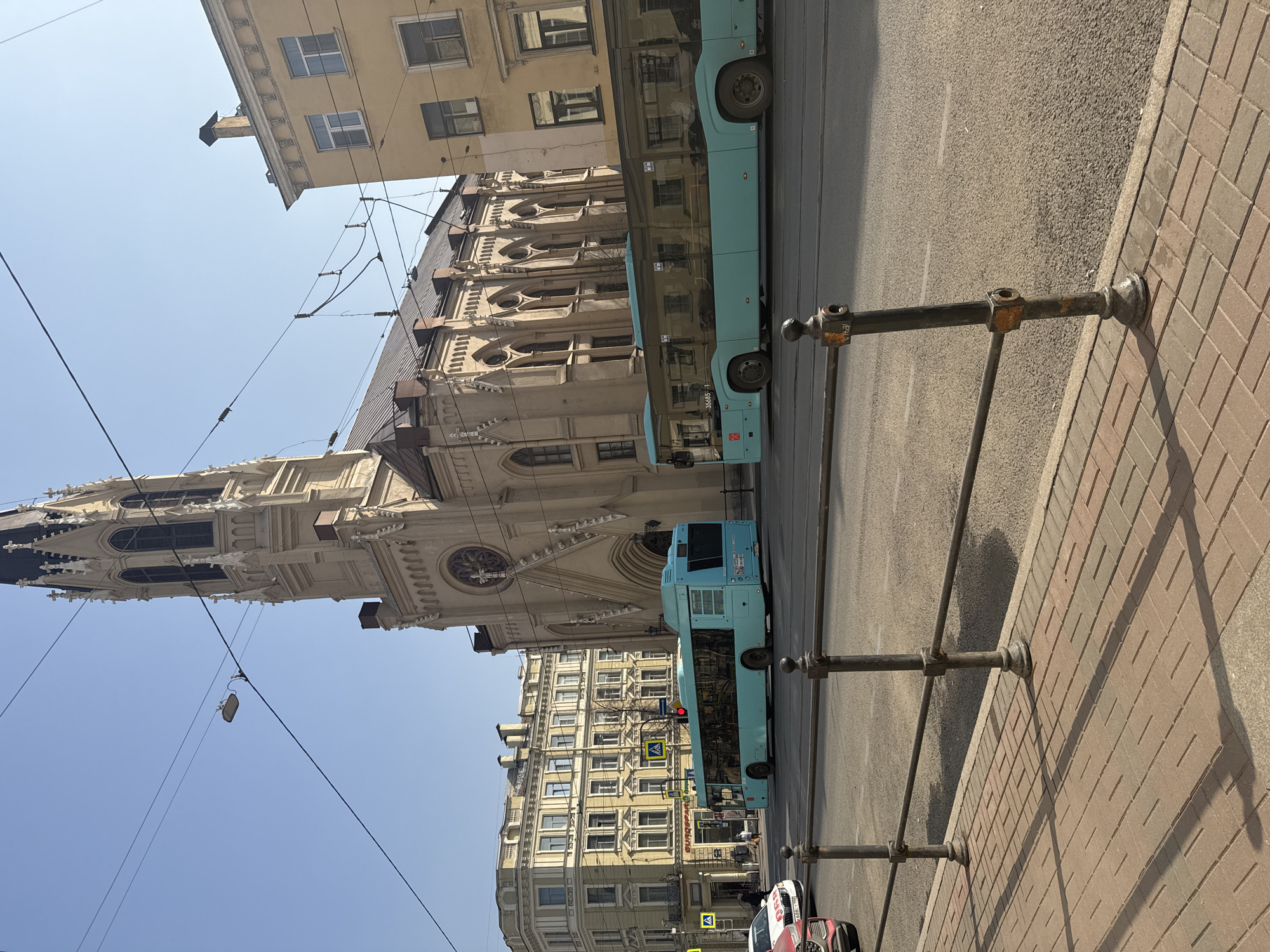 The image depicts a street scene in Saint Petersburg, Russia. Dominating the left side of the frame is St. Peter's Church, an example of neo-Gothic architecture. The church's spire rises prominently against a clear blue sky. Adjacent to the church are several buildings with classical architectural details, including windows, cornices, and ornamented facades. Two public buses painted a light blue color are present, traveling along the road that separates the buildings from the sidewalk. The camera appears to have been positioned on the sidewalk, where a metal railing runs along its edge. The sidewalk is paved with rectangular bricks. The presence of overhead power lines is also visible. A few pedestrians are partially visible, near the crosswalk, and a car can be seen further down the road.