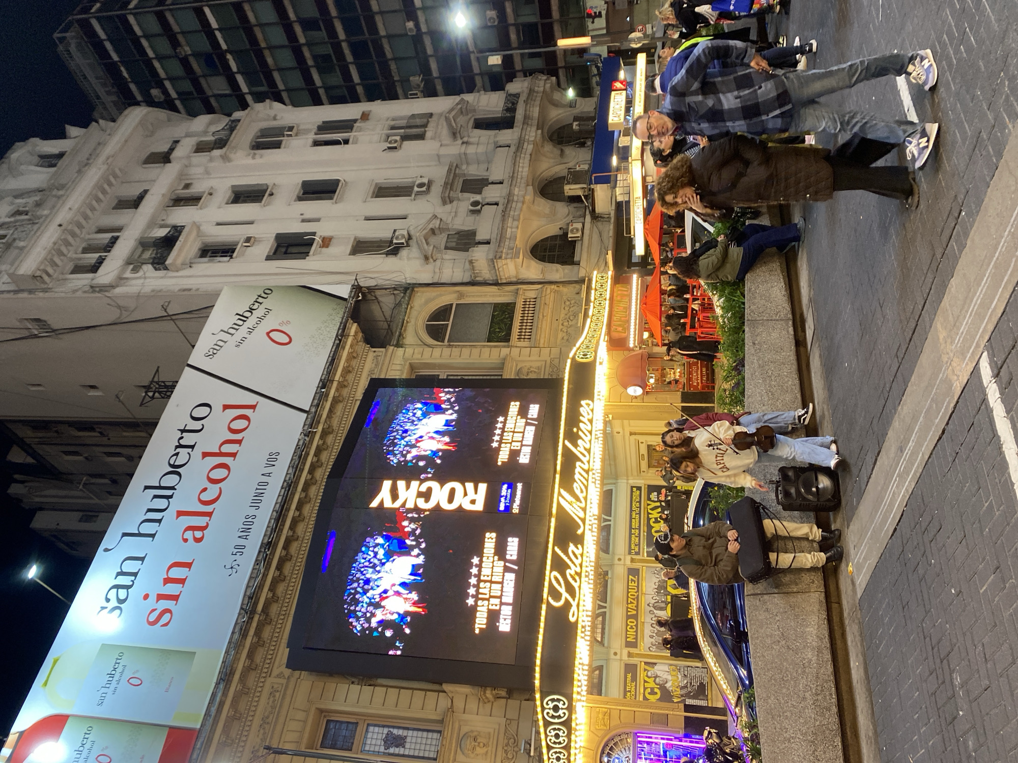The image depicts a street scene at night in Buenos Aires, Argentina, in front of the Lola Membrives theatre. The theatre's marquee is lit up, advertising the musical "Rocky," and the building has decorative architectural features. A large billboard advertises "San Huberto Sin Alcohol."
Several people are visible. A man and woman stand on the sidewalk, seemingly observing the theatre. Further down, a group of three people, including at least one person holding a violin, are sitting on the low wall. There is also a man sitting on the wall, holding a violin case.
The environment includes the theatre building, surrounding buildings with windows and air conditioning units, street lights, and the paved sidewalk. A restaurant/bar is visible next to the theatre. The scene suggests a lively urban setting with evening entertainment.