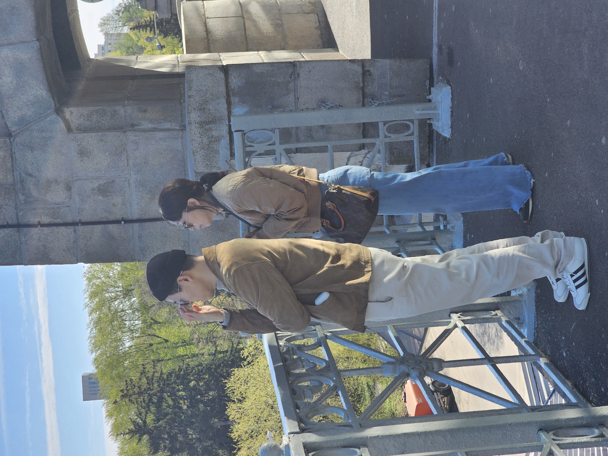 Two people are standing on a bridge in Moscow, Russia, looking over the railing. The man is wearing a black beanie, glasses, a brown jacket, and white pants with Adidas shoes. The woman is wearing glasses, a brown jacket, and blue jeans with a brown purse. They are standing on a concrete bridge with a gray metal railing. There are trees and buildings in the background. The sky is blue with some clouds.