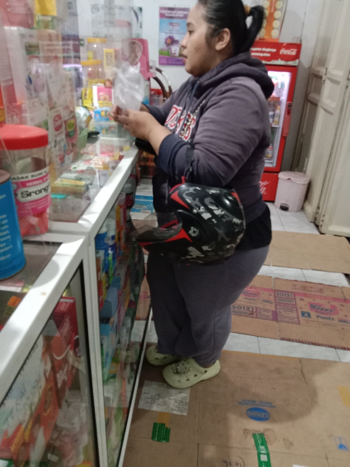 A woman is depicted standing inside a retail establishment in Turunsih, Indonesia. She wears a dark hooded sweatshirt, dark pants, and light green clogs, with her dark hair tied back in a ponytail. A black, red, and camouflage pattern motorcycle helmet is held under her left arm. Her hands are engaged with a translucent plastic bag. She stands facing a white-framed glass display counter filled with various packaged products. On the counter's left side, a blue container and a red-lidded jar branded "Srong" are visible. The tiled floor is partially covered by cardboard sheets, some bearing text such as "50121" and "Happy Pants." In the background, a red Coca-Cola branded refrigerator and a light pink waste bin are positioned against a light-colored wall.