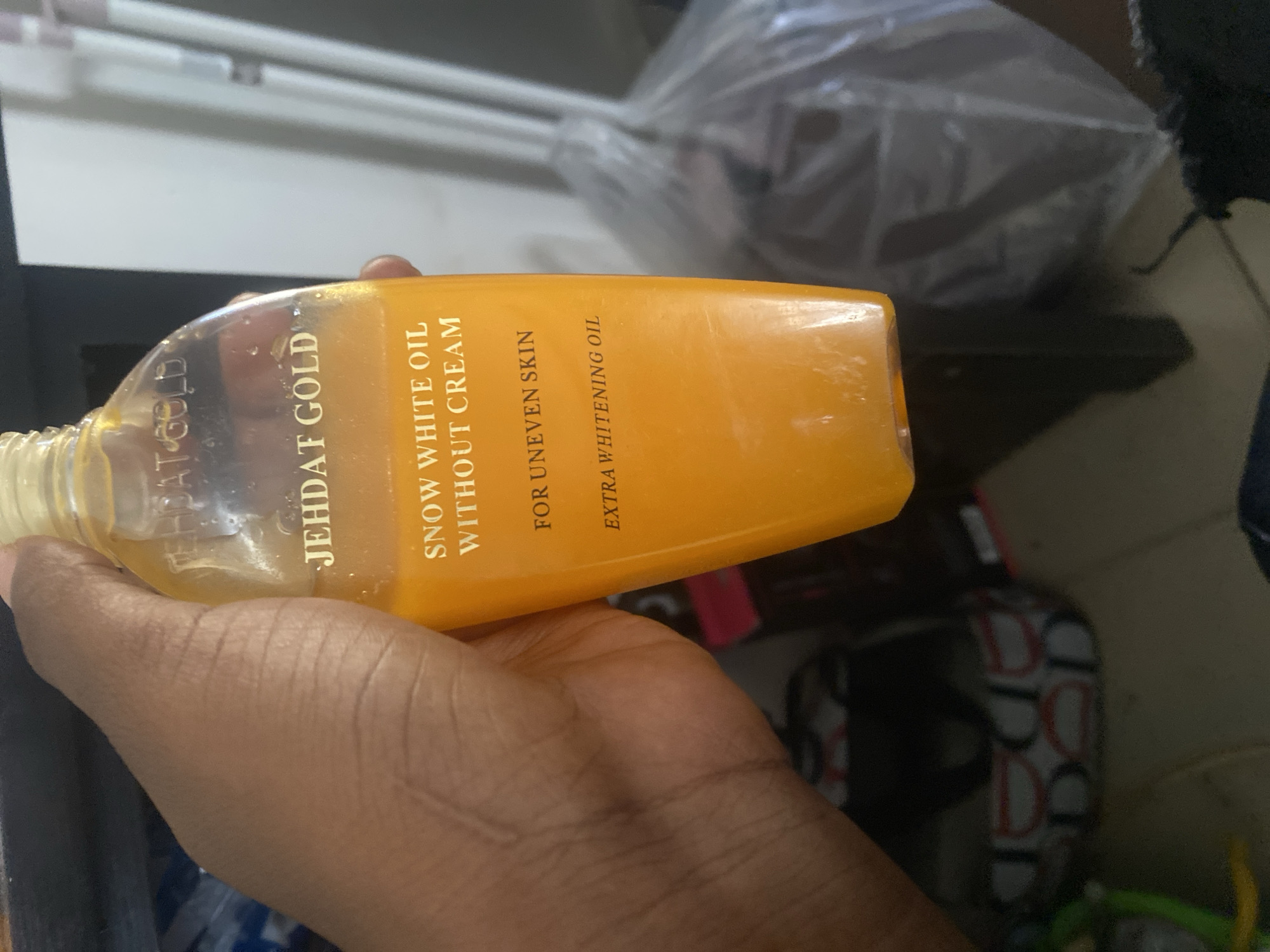 In Kubwa, Nigeria, a person's hand is holding a clear plastic bottle filled with a bright orange liquid. The bottle has white text on it that reads "JEHDAF GOLD", "SNOW WHITE OIL WITHOUT CREAM", "FOR UNEVEN SKIN", and "EXTRA WHITENING OIL". The background is blurred and appears to be indoors, with some shelving and a large plastic bag visible.