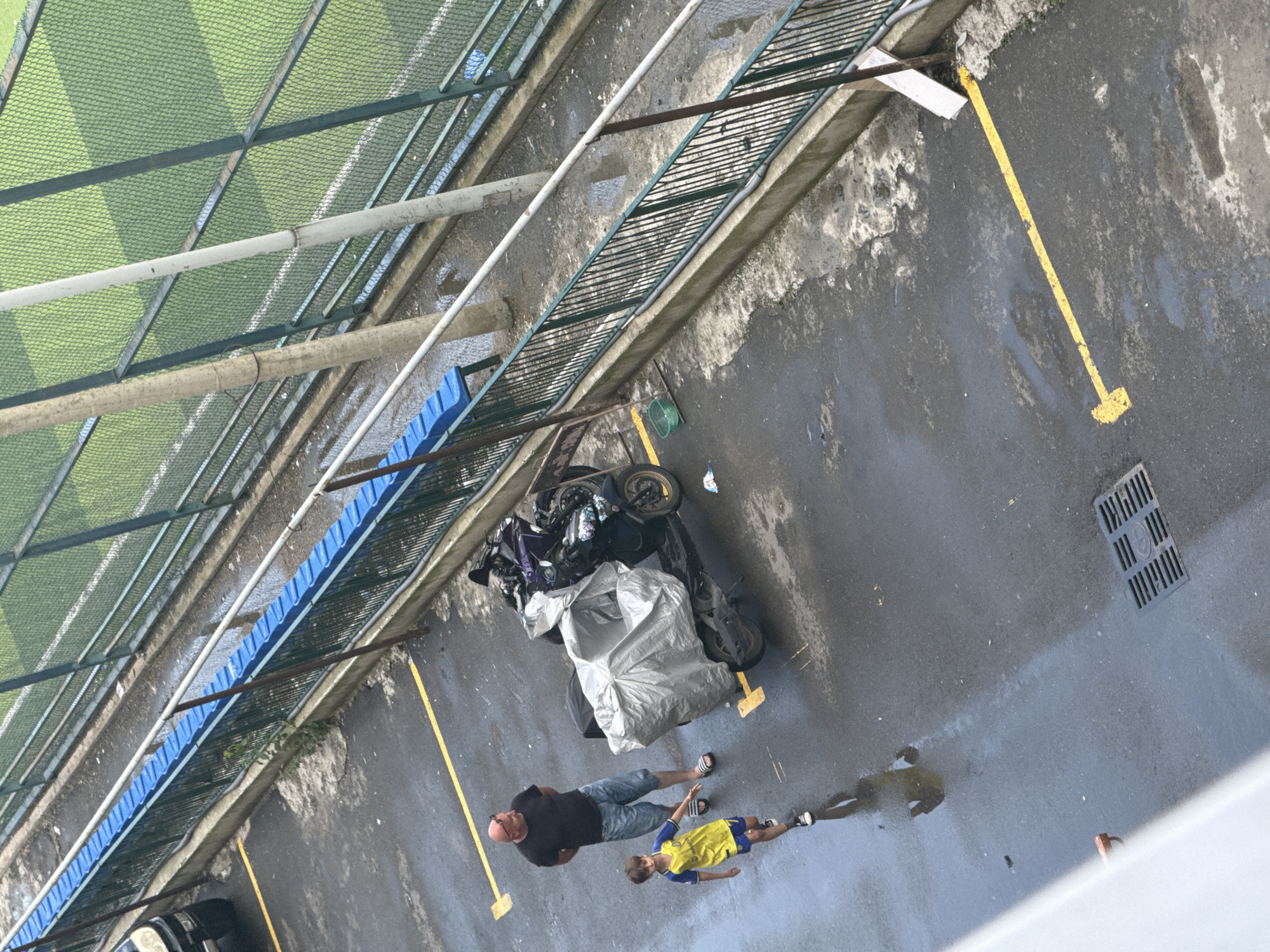 An overhead, eye-level shot captures an outdoor scene with a man and a boy standing on wet asphalt. The man, bald with sunglasses perched on his head, wears a black t-shirt and denim shorts. The boy, in a yellow and blue soccer jersey and shorts, walks toward the man. They are near a scooter covered with a gray tarp, with another dark scooter partially visible behind it. Yellow parking lines are marked on the asphalt. To the right of the scooters, a green fence with blue seating is visible, separating the asphalt from a green field with white markings, likely a sports field. Several concrete poles and a metal fence run along the edge of the field. The ground is wet and reflective, suggesting recent rain. A drain grate is present in the lower right portion of the image.