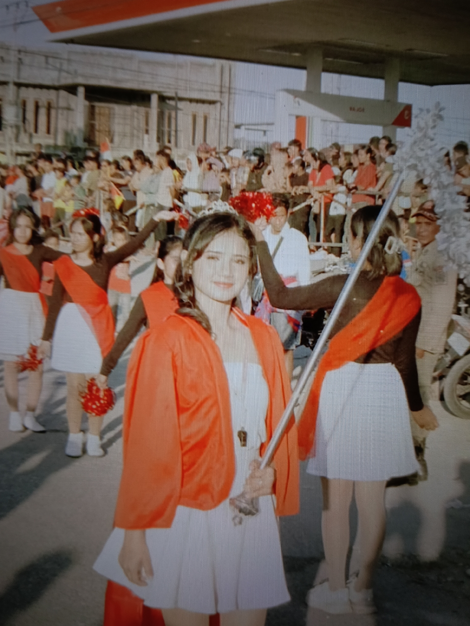 This image depicts a street scene in Raha, Indonesia, likely during a parade or procession. In the foreground, a young woman wearing a white dress and an orange jacket with a sash smiles at the camera, holding a metallic rod. Behind her, several other young women, dressed in similar uniforms consisting of white skirts and dark tops with red sashes, are marching. They are also holding red pom-poms. A crowd of spectators lines the street, some visible in the background, observing the procession. Above the crowd, a structure resembling a gas station canopy is visible, with signage including "RAHA" which indicates the location. The overall atmosphere appears festive.