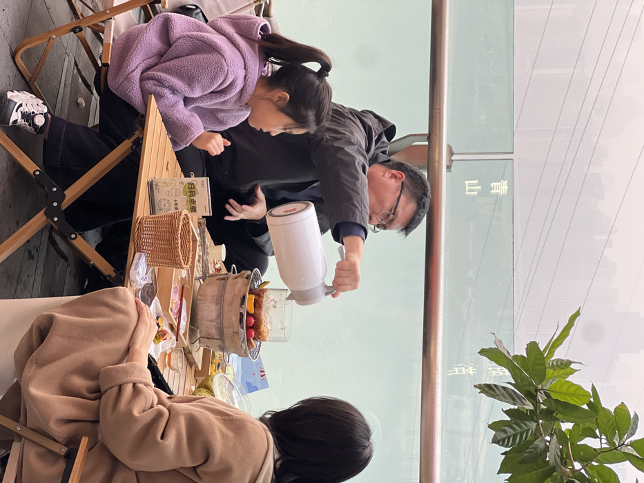 An outdoor scene in Yuzhong District, China, depicts three individuals engaged in a tea service alongside a body of water. An adult male, wearing glasses and a dark jacket, is centrally positioned, actively pouring liquid from a white electric kettle into a clear glass pot. The pot, containing sliced fruit and liquid, rests on a small, cylindrical charcoal stove. To his left, a young female child with dark hair tied in a ponytail, dressed in a purple fleece hoodie, observes the pouring action. To the male's right, an adult female in a light brown long coat is seated with her back partially to the viewer, also observing the table. The group is seated at a wooden folding table set with a small wicker basket, a white sign displaying "扫码点单" (scan code to order) and "6号桌" (Table No. 6), and assorted small dishes containing food items. Behind the table, a glass barrier, featuring the Chinese characters "青山" (Qingshan) and a partially visible "渝" (Yu), separates the seating area from a river. A hazy urban skyline and overhead power lines are visible in the distant background, with green tree foliage appearing in the upper right. The ground beneath the table is a dark grey surface.