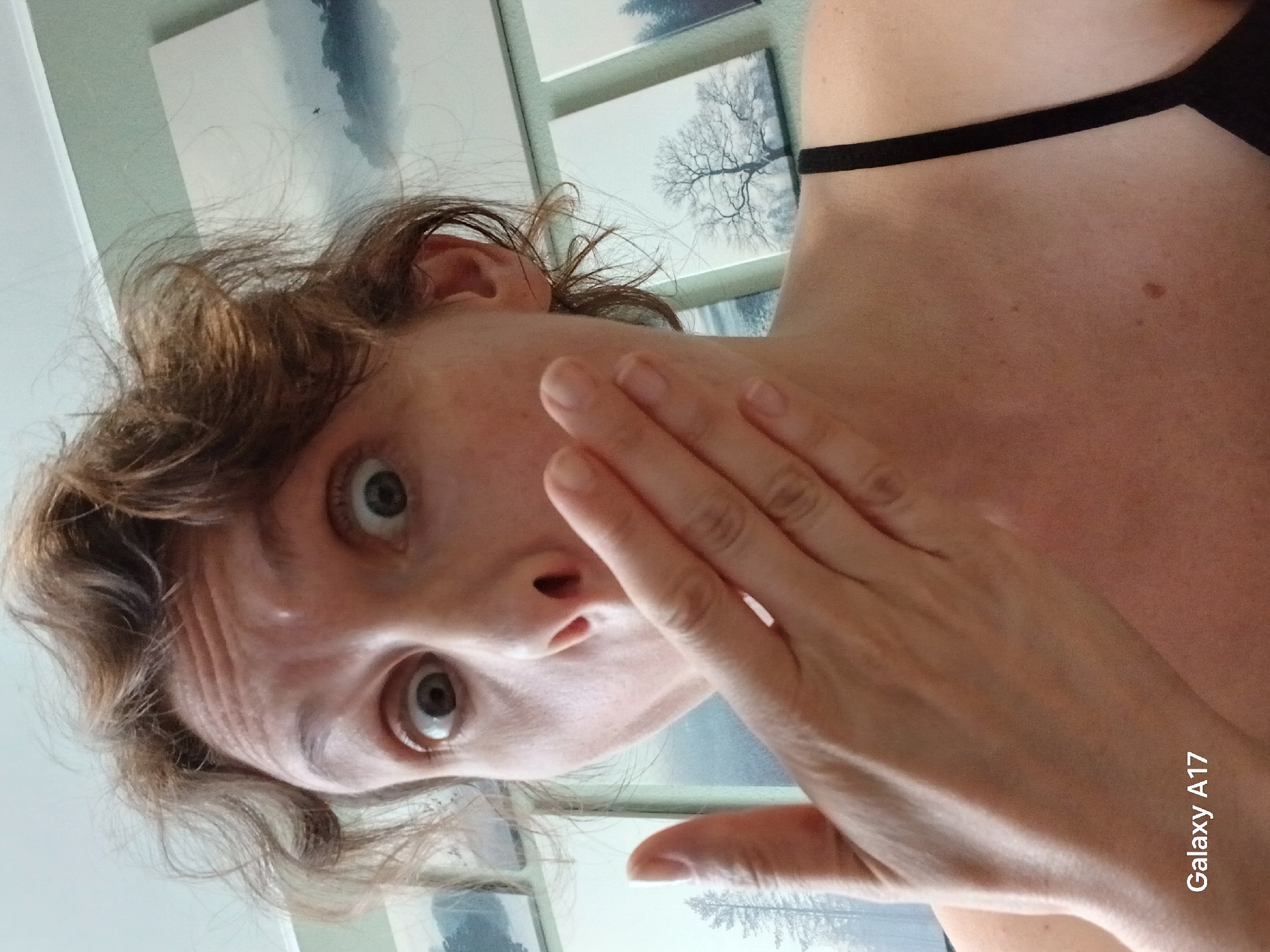 A selfie shows a woman with light skin and brown hair. Her eyes are wide and her mouth is slightly open in a surprised expression. Her right hand is raised to her face, fingers spread. She is wearing a black strap top. Behind her are several framed pictures of nature scenes. The bottom right corner of the image contains the text "Galaxy A17". The location is not Yekaterinburg, Russia.