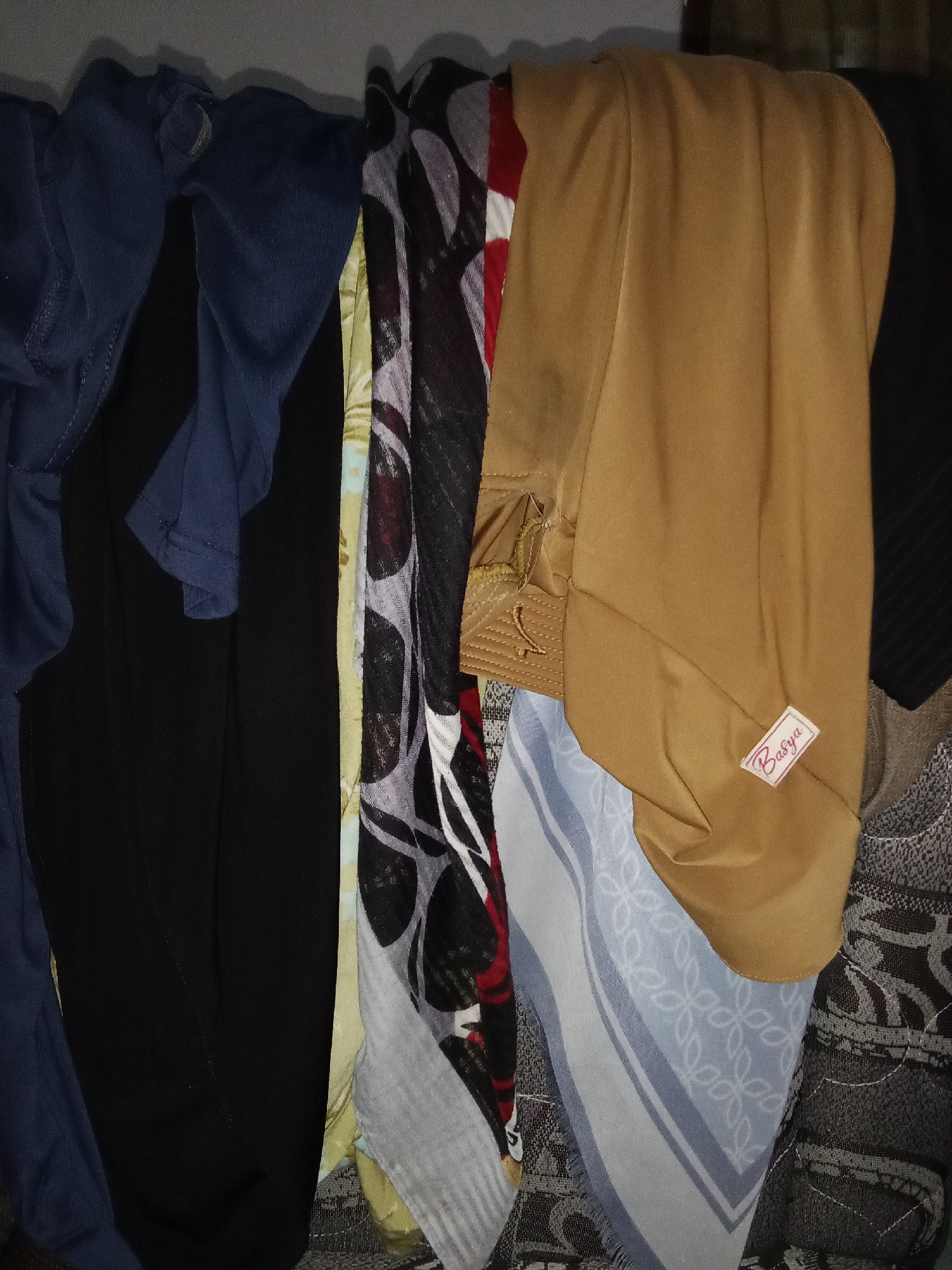 In Selowogo, Indonesia, this image captures a close-up of several pieces of clothing, possibly laundry, hanging on a line. The items are layered and close together, creating a textured visual. On the left, a dark blue fabric is visible, followed by a black garment. Next, a patterned fabric with black, white, and red designs is draped, and a lighter colored fabric with a yellow hue is partially visible behind it. A prominent, mustard-yellow garment occupies the central and right portions of the image, displaying a visible label that reads "Basya" in a stylized font. To the right of the yellow garment, a light blue and white patterned scarf with a fringed edge is hanging. The background is dark and indistinct, suggesting an indoor or shaded outdoor setting. The lighting is somewhat dim, with shadows playing across the fabrics.
