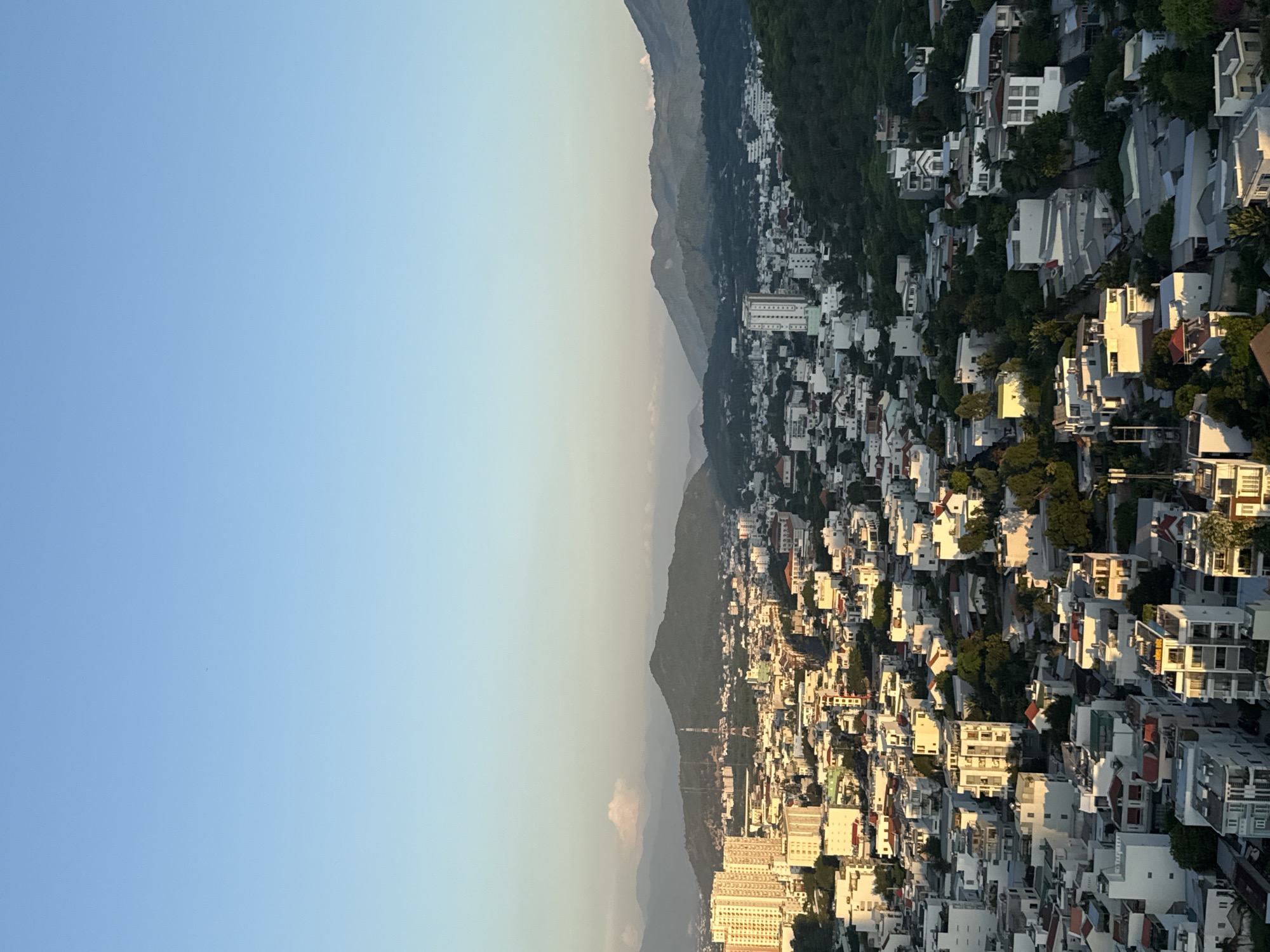 This is a high-angle, full-frame, vertical shot of the city of Nha Trang, Vietnam, taken at sunset. The sky is a clear, soft blue with a gradient to lighter shades near the horizon. In the foreground, a densely packed urban area is visible, characterized by a mix of low-rise buildings and taller apartment complexes. The buildings are predominantly white with some exhibiting red-tiled roofs. The setting sun casts a warm, golden light on the western-facing sides of the structures, creating stark contrasts between light and shadow. Lush green trees are interspersed throughout the city, particularly on the right side of the image, and appear to be climbing the foothills. In the background, rolling, tree-covered mountains form a natural backdrop, with layered mountain ranges fading into the distance. A prominent radio tower stands out against the landscape in the mid-ground. The scene depicts a vibrant city nestled against a natural landscape during the tranquil hours of sunset.