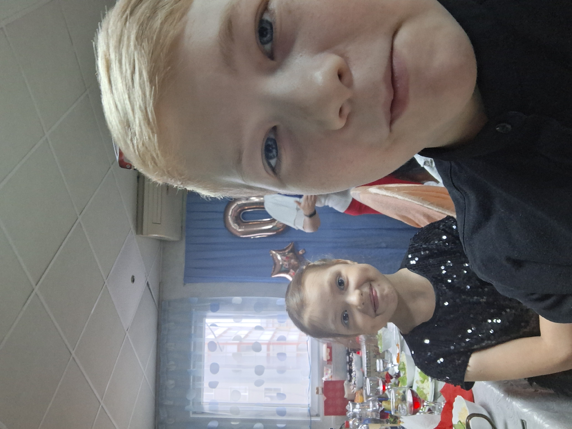 Two children, likely siblings, pose for a selfie in a room decorated for a birthday party, potentially located in Chaikovsky, Russia. The elder child, a blond boy, is in the foreground, wearing a black shirt. The younger child, a girl, is in the midground, wearing a black sequined shirt. A table laden with food and drinks is partially visible in front of her. Behind the children, blue curtains with a polka dot design hang in front of a window. Above the curtains, a gold "0" balloon and a star-shaped balloon suggest it may be a tenth birthday. Part of a person wearing a white shirt and red jacket is also visible. The room has a white tiled ceiling with a vent or air conditioning unit attached.