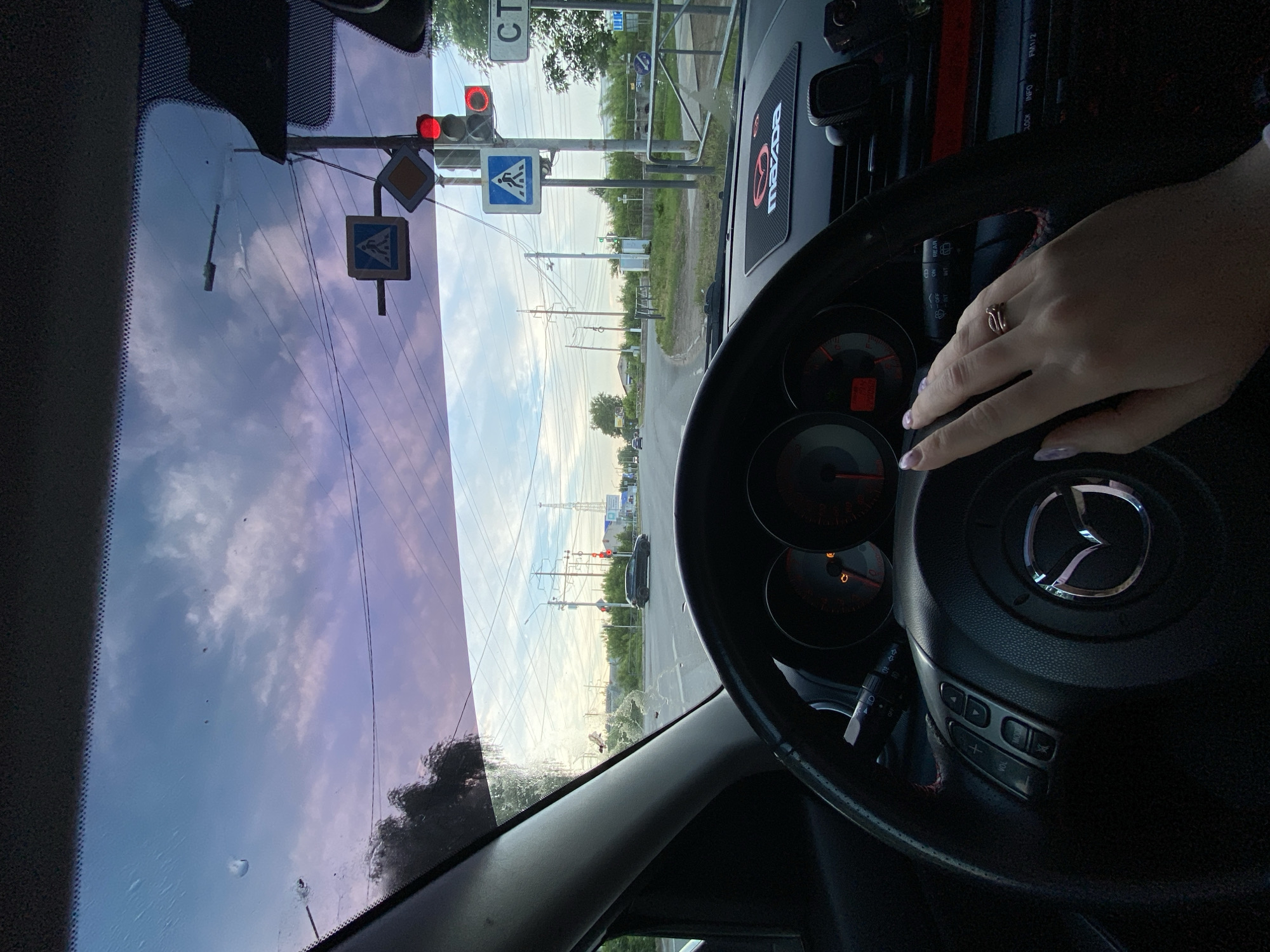 A view from inside a Mazda car at a red traffic light. The intersection features a pedestrian crossing sign and other road signage in Russian. Power lines are visible extending down the street. Based on the "CT" sign on a building in the background, the location is likely in the city of Chaikovsky, Russia. A hand wearing a ring rests on the steering wheel. The car's dashboard and controls are visible.