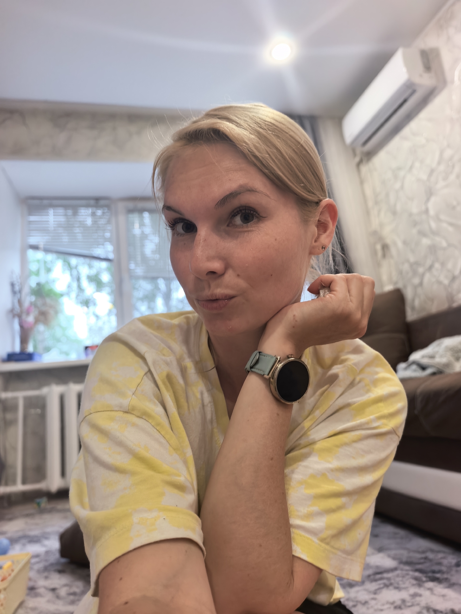 The image is a medium shot of a young, blonde woman with fair skin, looking directly at the camera with a slightly puckered expression. She is wearing a yellow and white tie-dye t-shirt and a smartwatch with a light blue band on her left wrist. Her left arm is bent, with her hand resting on her chin, and her right arm is resting on her lap. The background appears to be an indoor setting, possibly a living room, with a window covered by blinds to the left, a radiator underneath, and an air conditioning unit mounted high on the wall to the right. A brown sofa is visible in the far right of the frame. The floor is covered with a patterned rug. There is no specific indication in the image that it was taken in Sterlitamak, Russia.