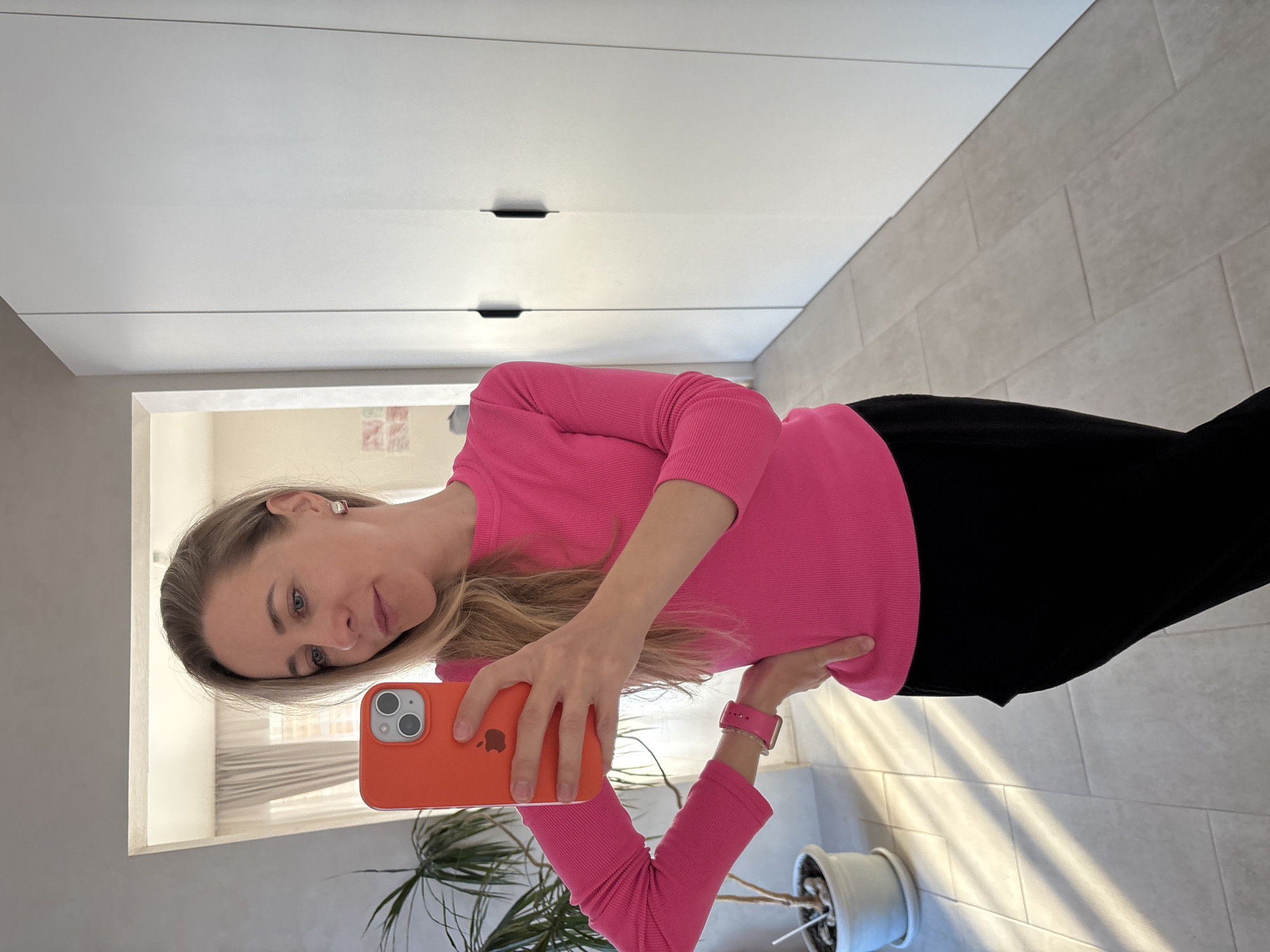A woman is taking a selfie with an orange-cased iPhone. She's wearing a pink ribbed top and black pants. The location appears to be indoors, possibly in an apartment or home, with a potted plant and white storage units visible in the background. It's not possible to confirm the specific city or country (Chaikovsky, Russia) based solely on the image content.