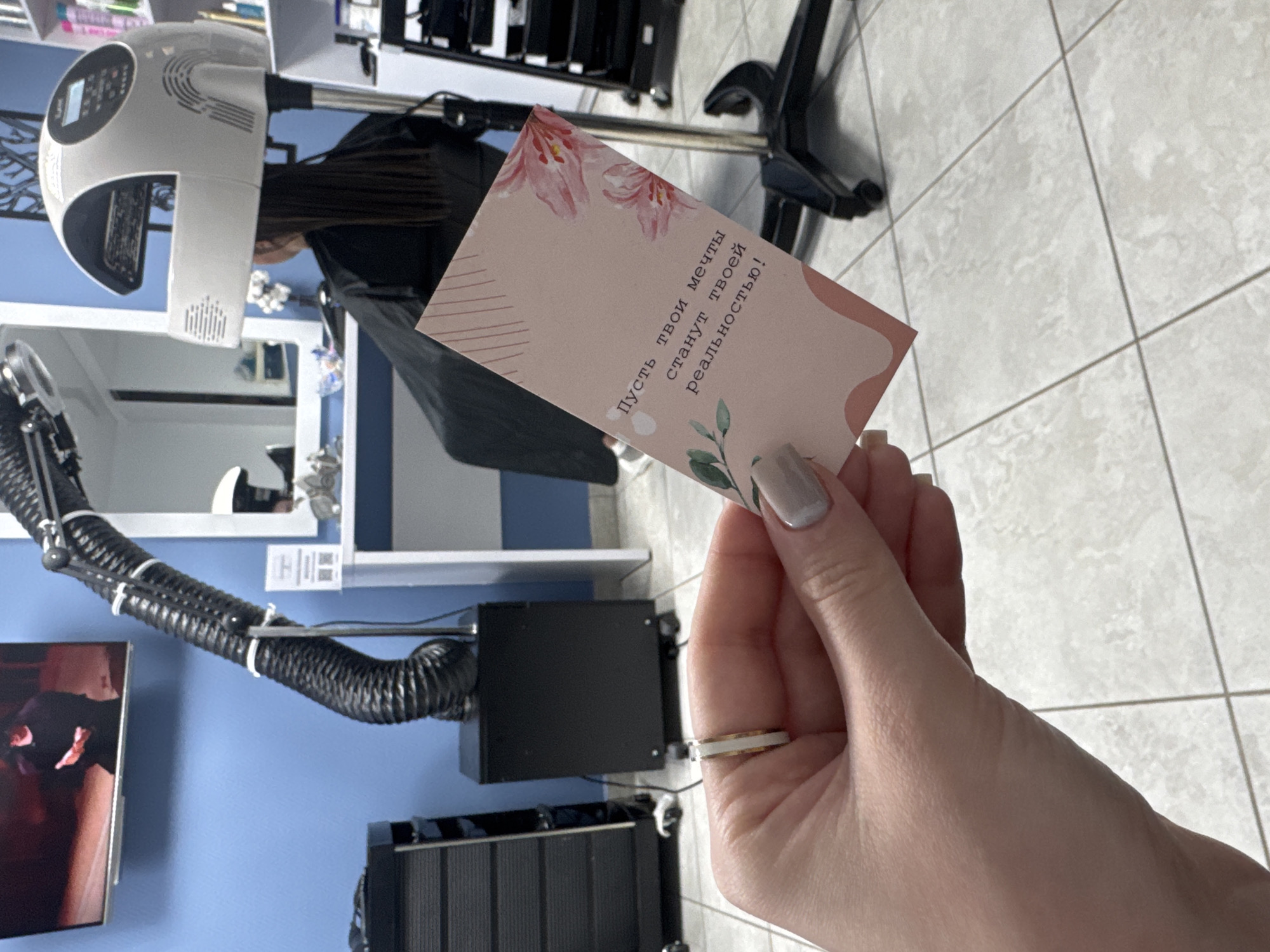 A hand is holding a pink card with Russian text in a hair salon. In the background, a hooded hair dryer is positioned over a client's head. The walls are painted blue, and the salon features tiled floors, salon chairs, and other salon equipment. The city is Chaikovsky, Russia.