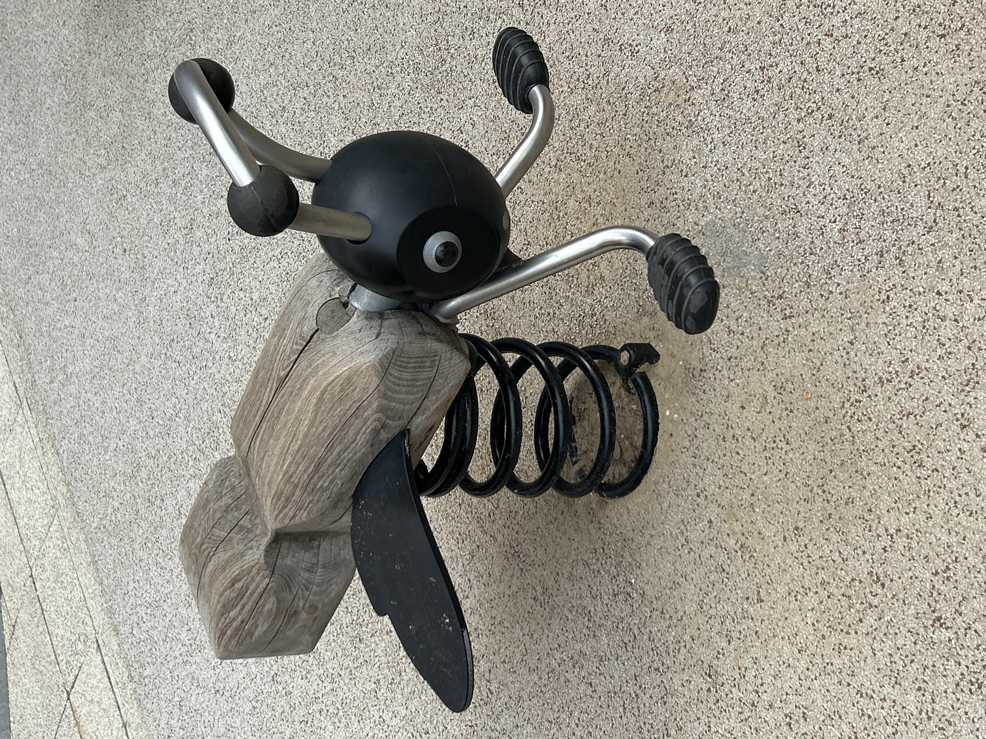 A spring rider toy, designed to resemble an insect, is situated outdoors on a speckled, beige-colored surface. The toy features a wooden seat that resembles the insect's body, with a black wing attached to its left side. Beneath the seat, a black coil spring connects it to a base. The front of the toy is a black, round head with a single grey eye and two silver antennae topped with black spheres. The insect has two sets of metallic legs, each ending in a black footrest. The overall scene depicts a playground element in an urban outdoor setting, specifically located in the city of Khimki, Russia.