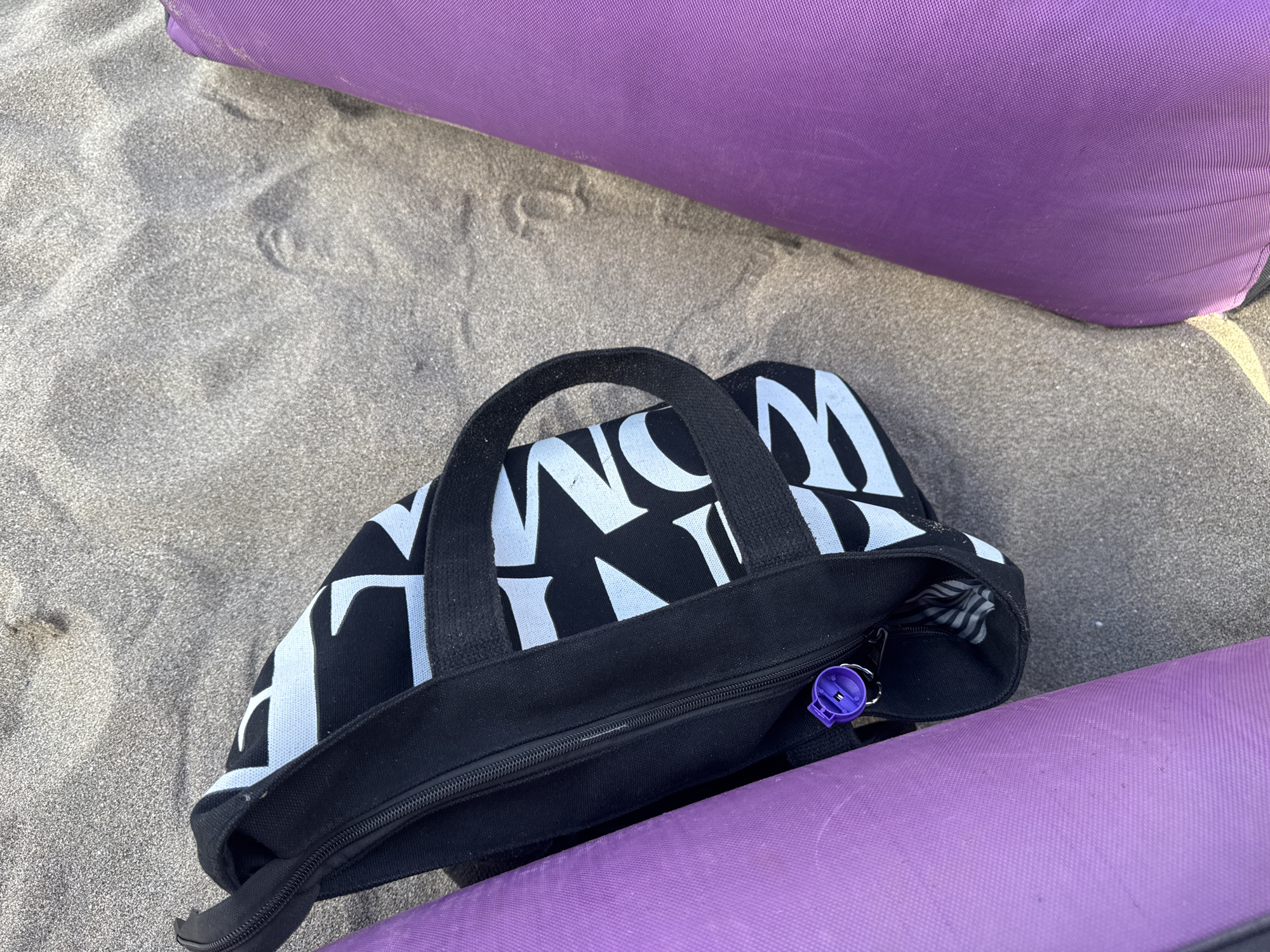 The image shows a black tote bag with white lettering placed on sand. Two purple yoga mats are positioned on either side of the tote bag. The bag has a zipper closure, and a purple object, possibly a key fob or bottle opener, is attached to the zipper pull. The sand appears slightly damp and shows faint imprints. The setting is likely Kuta, Indonesia, given the tropical context often associated with beach and yoga activities in that region.