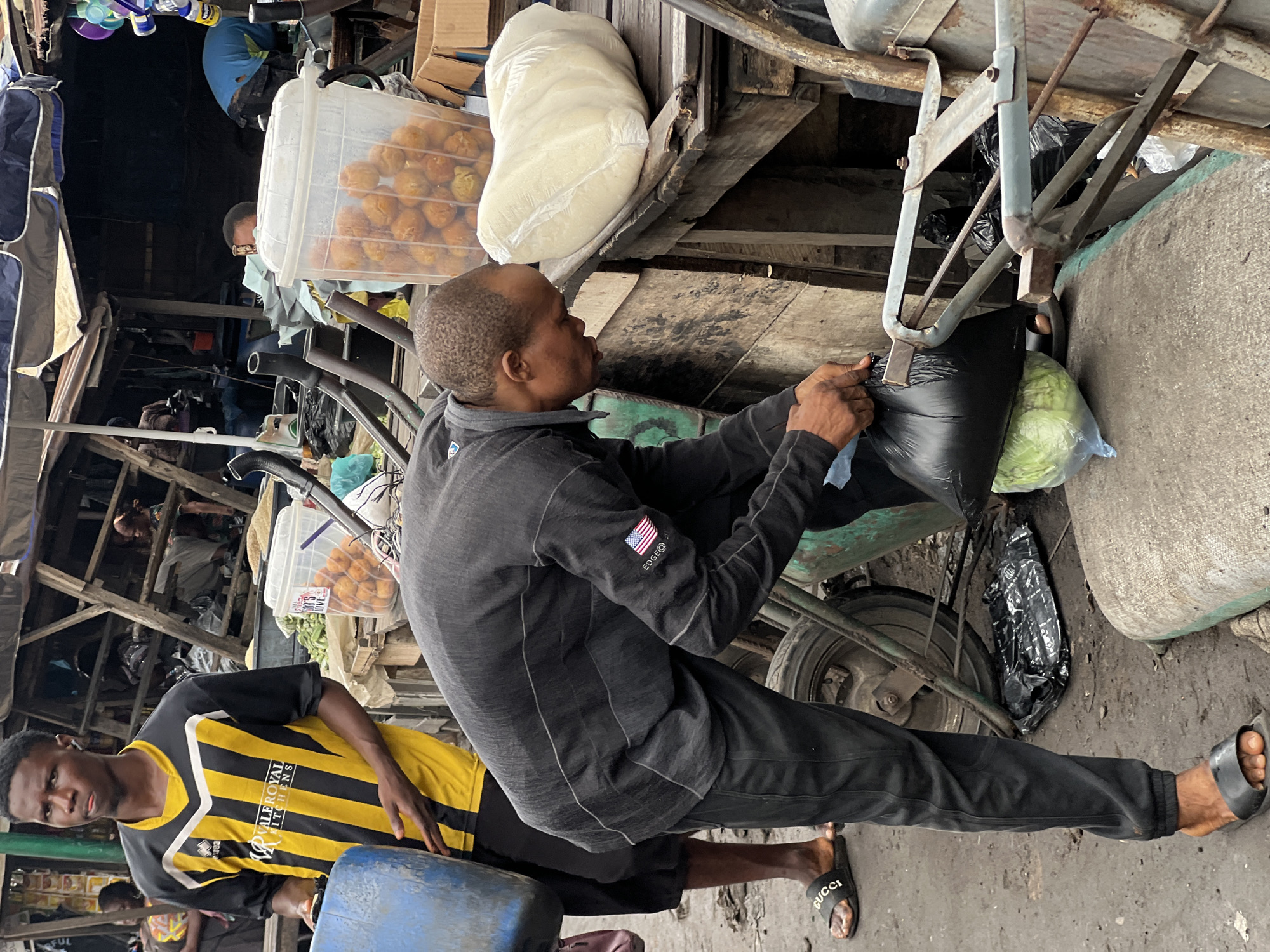 In Nkpor, Nigeria, a man in a black sweater and pants bends down to pick up a black plastic bag from a wooden market stall. He wears black Gucci sandals. Beside him, another man in a yellow and black striped shirt holds a blue container. In the background, a wooden structure provides shade over market stalls displaying food items in plastic containers. The ground is dusty and appears to be a busy marketplace.