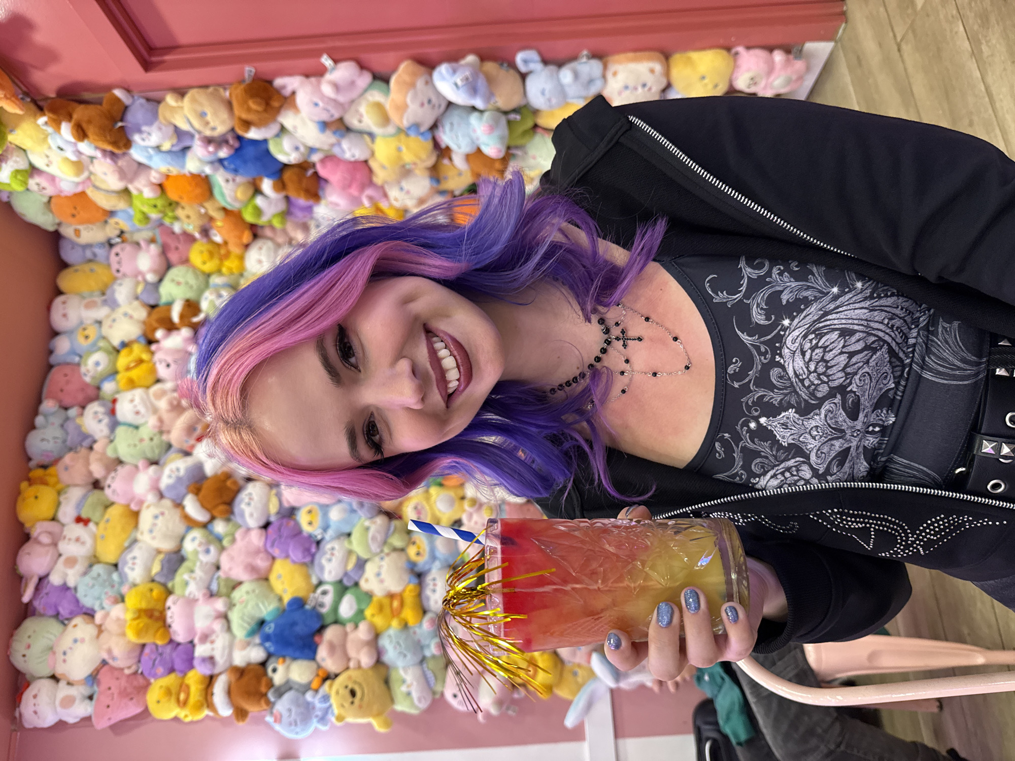 A smiling woman with purple and pink hair, wearing a black jacket and a silver and black top with a cross necklace, is holding a colorful drink in front of a wall filled with plush toys. The drink is in a textured glass and has a blue and white striped straw and gold tinsel. The background wall is a pastel pink and is covered in various small plush toys. The scene appears to be indoors. The location in the city of Madrid, Spain, cannot be confirmed based solely on the visual content of the image.
