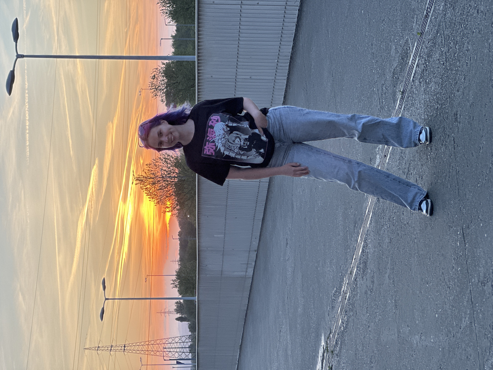 A young woman with purple hair stands on a gray asphalt surface next to a metal barrier, likely a bridge or elevated walkway. She wears a black t-shirt with a graphic print and light blue jeans. The setting sun casts a warm orange glow in the background, visible behind trees and utility poles. The sky is streaked with contrails. The location is plausibly within the City of Zagreb, Croatia, given the architectural and environmental context.