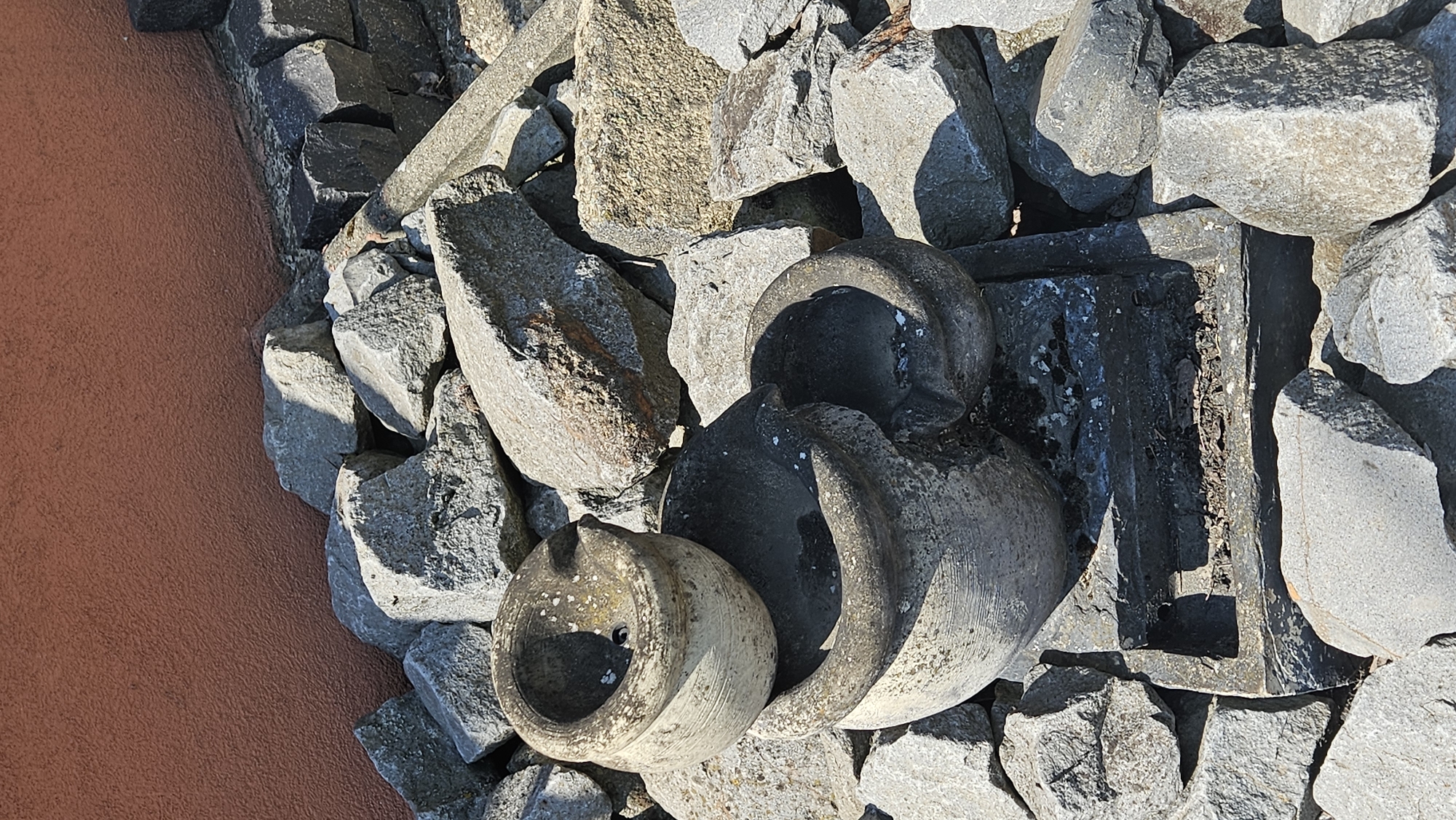 In Vyšné Nemecké, Slovakia, a natural outdoor scene unfolds against a textured, reddish-brown wall. The foreground is dominated by a collection of weathered grey stones of various sizes, creating a rough, uneven terrain. Nestled within these stones are three large, grey, bowl-like objects. The largest is positioned at the bottom, a broad, deep basin. Above it, slightly to the left and right, are two smaller, similarly shaped bowls, one resting atop the other. The surfaces of these bowls show signs of age and exposure to the elements, with some pitting and discoloration. A dark, rectangular base is visible beneath the largest bowl. The overall impression is one of an outdoor display or perhaps a decorative garden arrangement, characterized by natural materials and a rustic aesthetic.