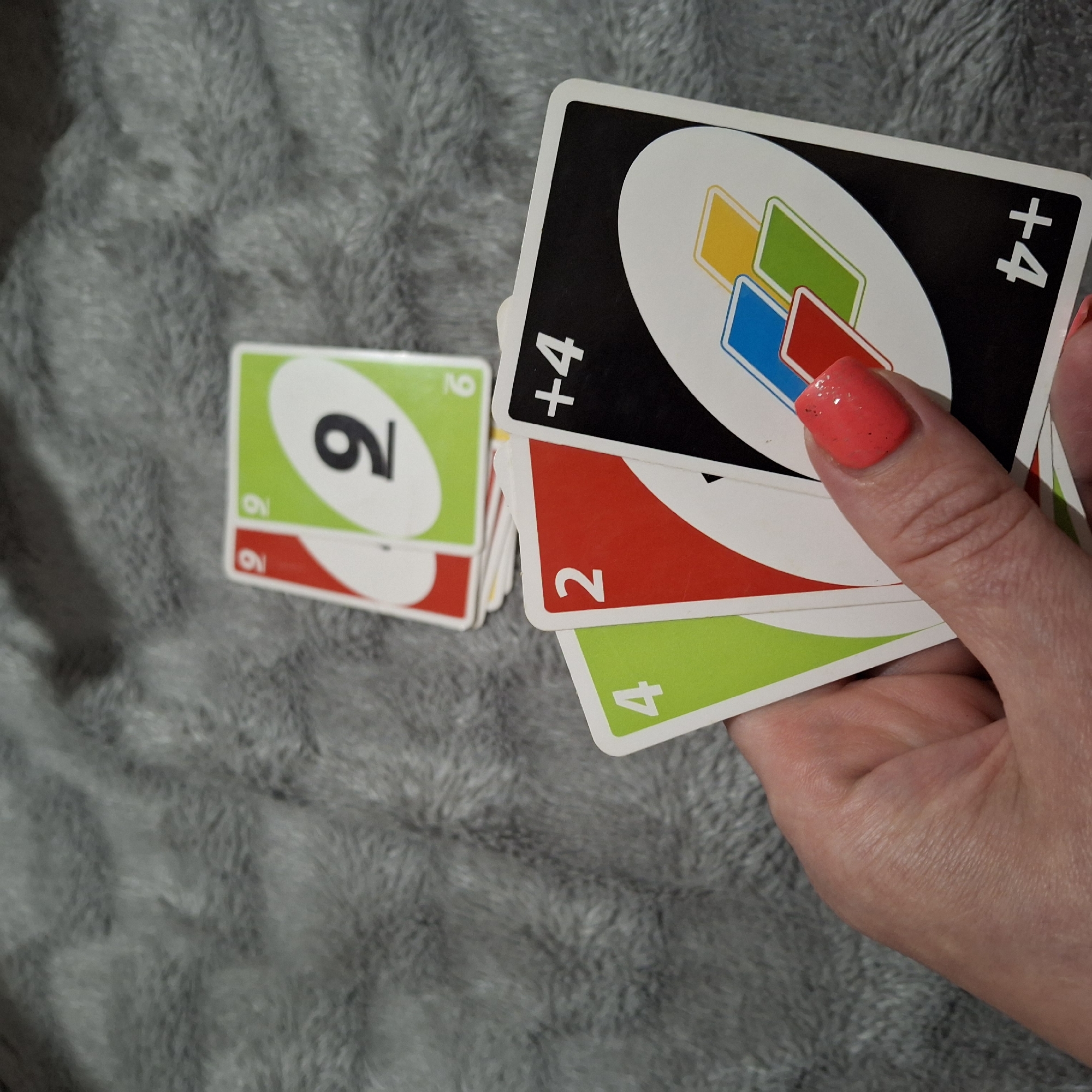 A person's hand, adorned with bright coral nail polish, holds a fan of UNO cards on a gray, fuzzy surface. The cards visible are a green '4', a red '2', and a black '+4' card. In the background, two more UNO cards, a red '9' and a green '9', are partially visible. The scene appears to be indoors, possibly on a couch or bed, given the texture of the surface. No specific location in the city of Unknown, Unknown can be determined.