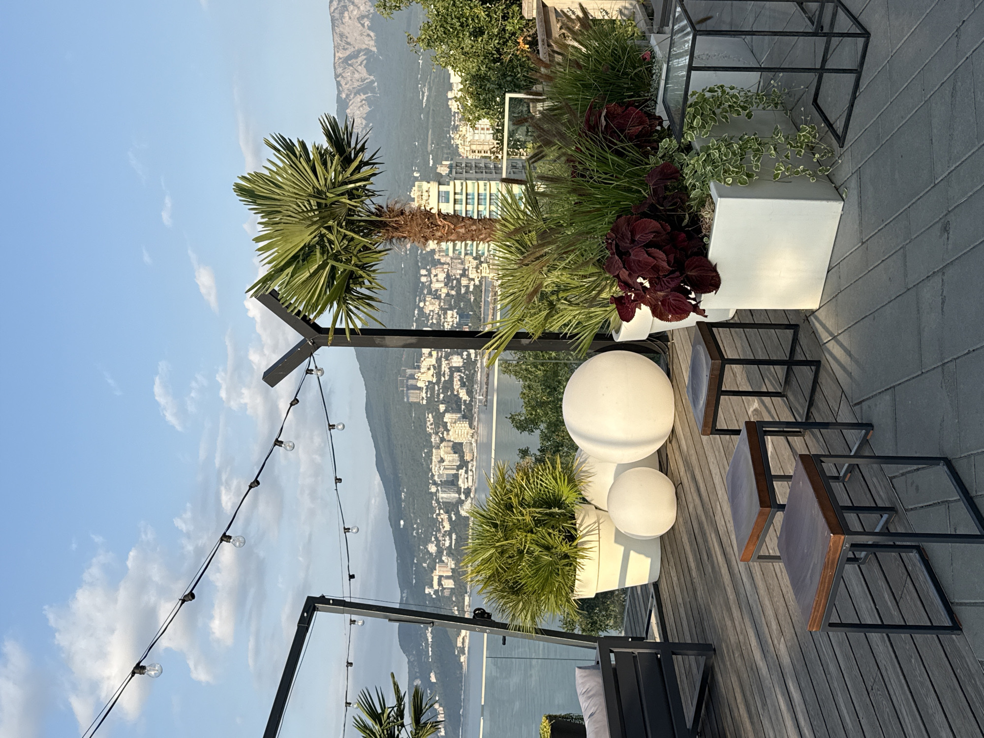An outdoor seating area with a city and sea view is captured. The foreground features a wooden deck with several dark-legged stools with wooden tops. To the right, a white square planter overflows with green plants, including cascading vines. Behind it, a dark metal framed table with a glass top stands. In the mid-ground, large white spherical planters are arranged on the deck. A prominent potted palm tree is situated to the right of these planters, with a backdrop of other greenery. Beyond the seating area, a body of water stretches out, meeting a city skyline featuring numerous buildings. Mountains form the distant horizon, with a clear blue sky and scattered clouds above. The scene appears to be set in Yalta, Ukraine, given the distinctive coastal city and mountainous landscape.
