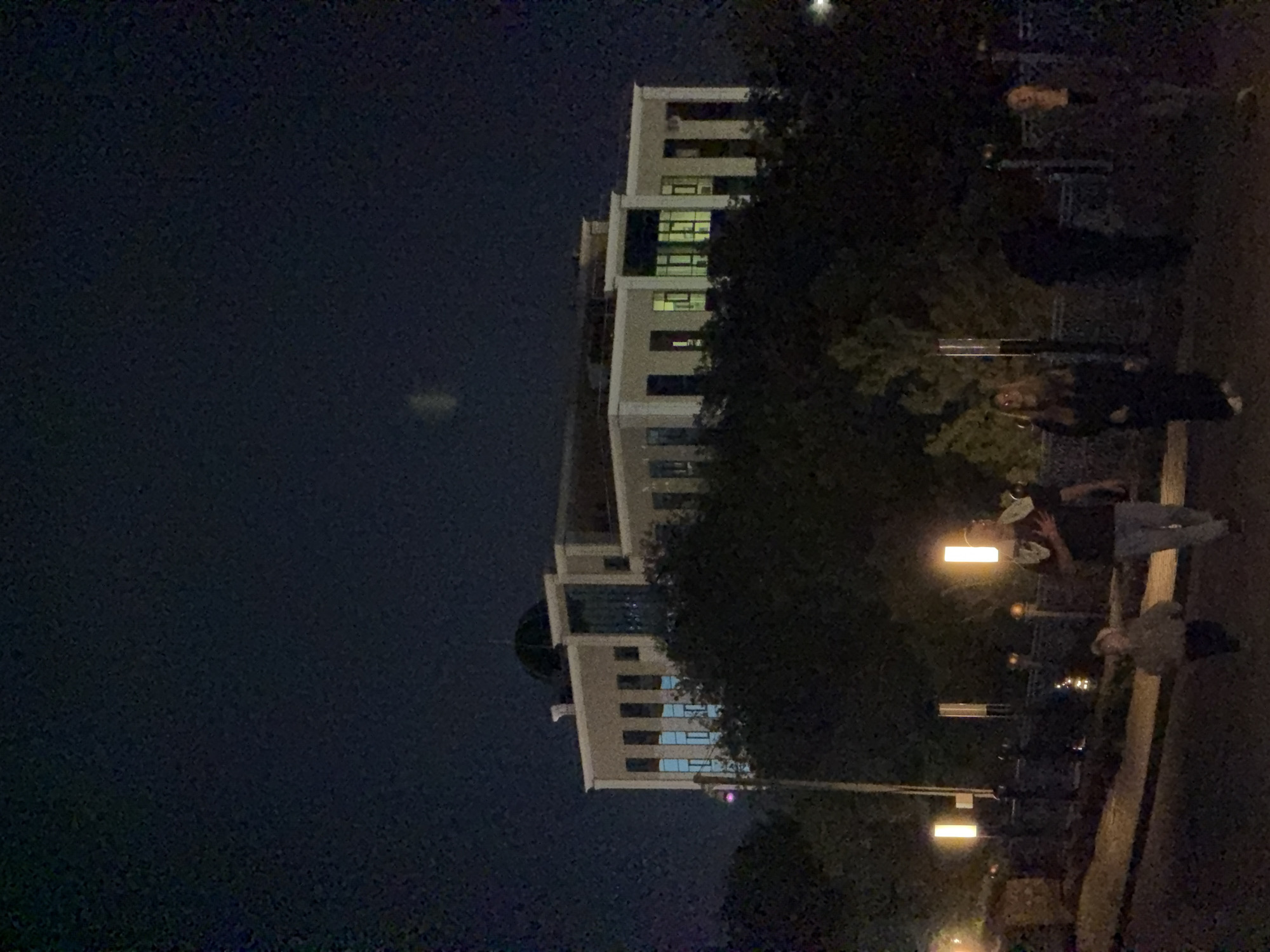 An outdoor, nighttime scene depicts a building with multiple illuminated windows, partially obscured by dark foliage. The building appears to be modern, with a light-colored facade and a domed roof section. In the foreground, several individuals are visible walking on a paved path. A child in a light-colored jacket walks ahead of an adult male wearing a dark shirt and light pants. Further to the right, two adult females are walking, one in a dark outfit and the other in a darker, possibly patterned garment. Several lampposts with tall, rectangular, illuminated fixtures line the path. The background is dark, suggesting a night sky. The location is identified as Pavlodar, Kazakhstan.