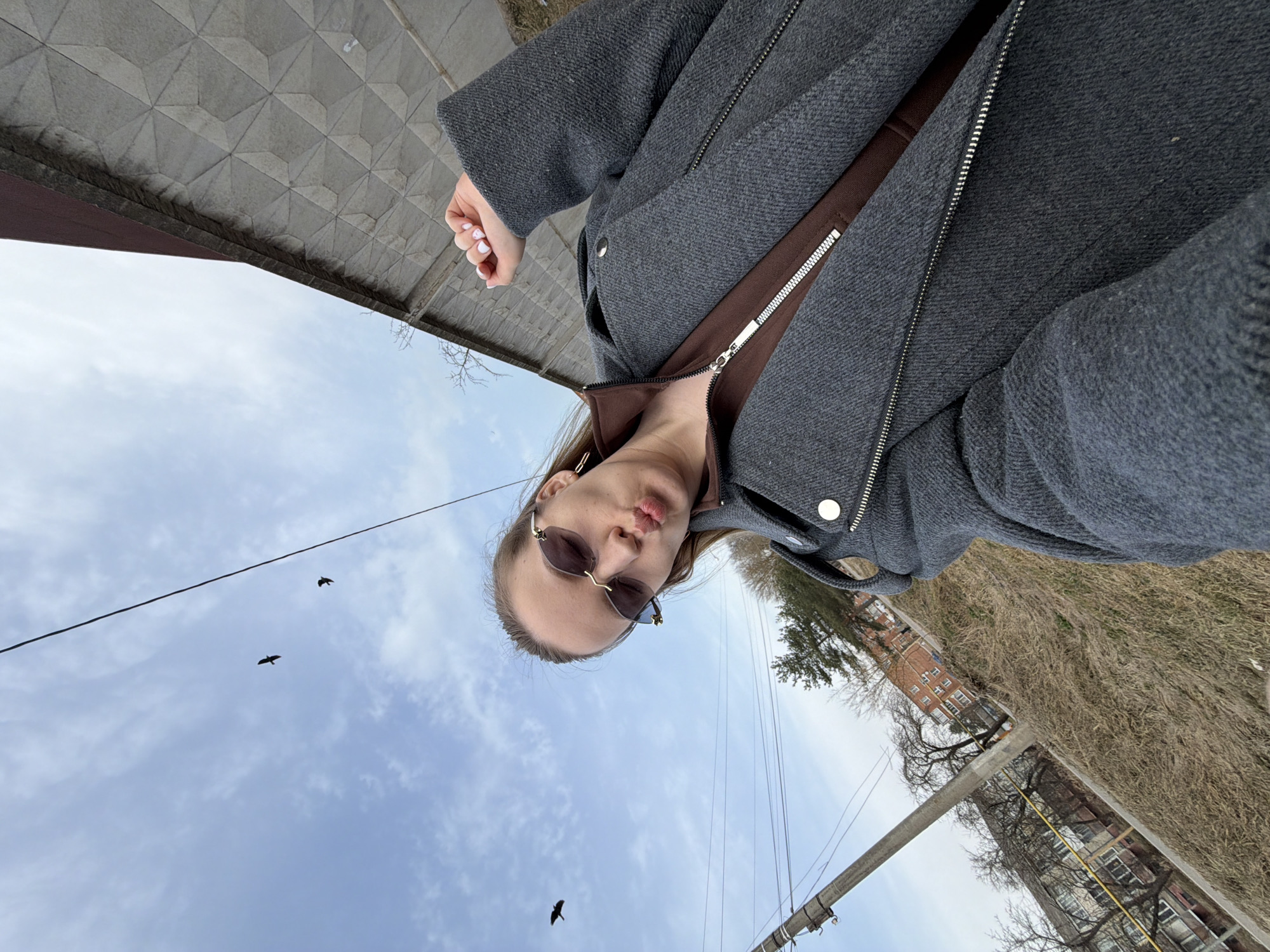 A person is visible in the image, turned so that the ground is above them and the sky is below, suggesting the image was rotated. The person is wearing sunglasses, a gray jacket with a brown lining, and earrings. Their lips are pursed. Overhead are electrical wires and a few birds in the sky. Buildings and trees are visible in the background, as well as some dry grass. The setting appears to be Reshetnikovo, Russia. The street is paved with textured gray slabs. The lighting suggests it's daytime.