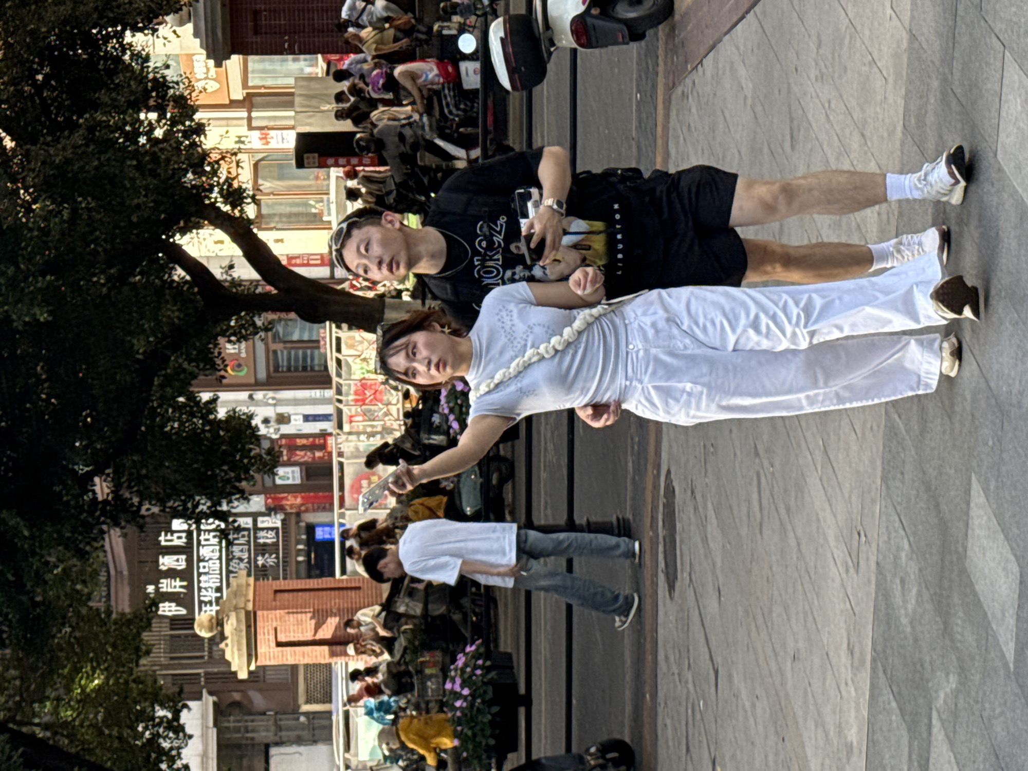 A street scene in Yuzhong District, Chongqing, China, shows a couple standing on a paved walkway. A woman in a white shirt and pants holds a cell phone in her right hand. Next to her is a man wearing a black t-shirt and black shorts. Behind them, other pedestrians walk along the street, with parked scooters and motorcycles visible. Buildings with Chinese signage line the street, partly obscured by a tree.