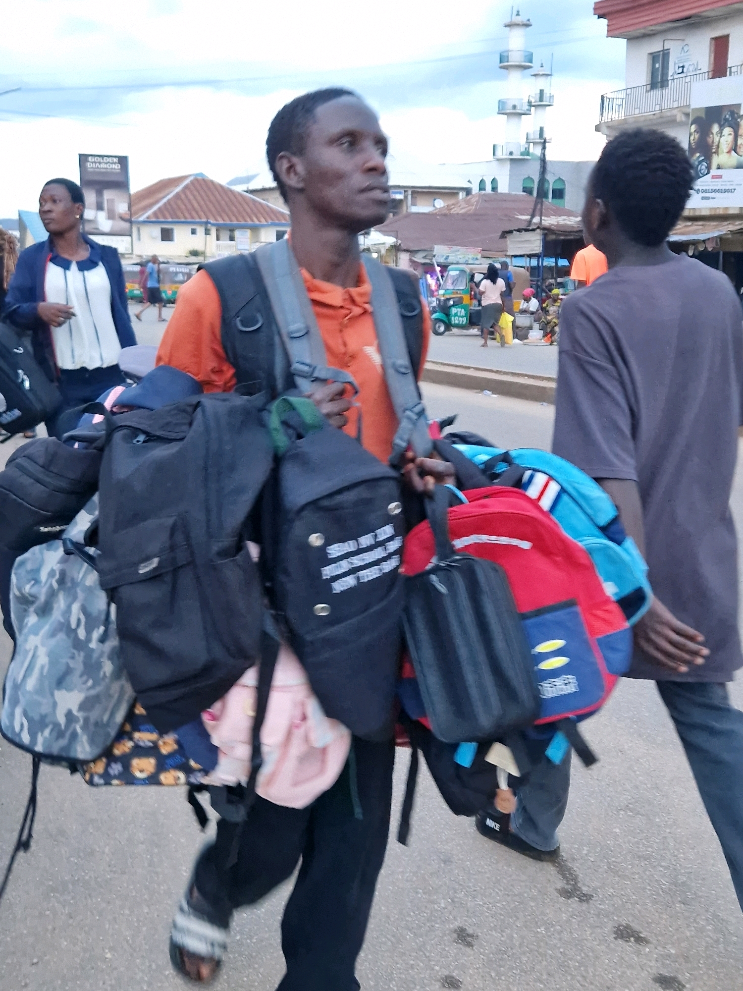 Two African men are walking on a paved road in what appears to be a busy street in Jos, Nigeria. The man in the foreground, wearing an orange shirt and a backpack, is carrying multiple bags, including a camouflage backpack and a red and blue backpack, in addition to his own. The man in the background, wearing a gray t-shirt, is also walking away from the camera. The street is lined with buildings, some of which appear to be shops, and there are other people and vehicles visible in the distance, suggesting a bustling urban environment. The sky is overcast with scattered clouds. The man in the foreground is wearing sandals with white and gray stripes, while the man in the background is wearing dark pants and what appear to be black sneakers with a white "NIKE" logo.