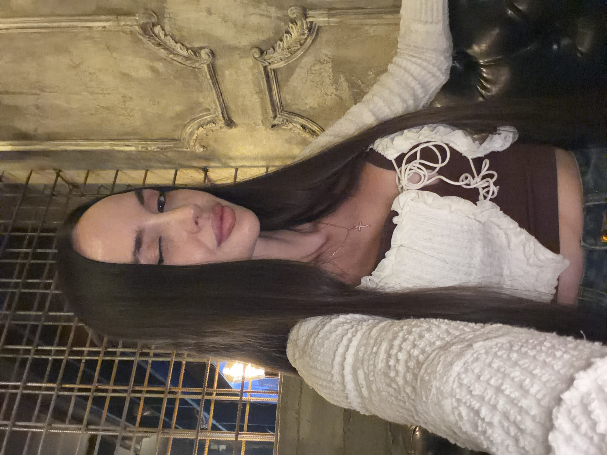 A young woman with long, dark hair is taking a selfie in what appears to be a dimly lit, ornate indoor space, possibly a restaurant or bar. She is wearing a white, textured cardigan over a dark brown top with a lace-up front. A delicate silver necklace with a cross pendant is visible. Her left eye is closed in a wink, and her lips are slightly parted. The background features a rustic, textured wall with decorative molding on the right, and a metal grate or window with lights visible through it on the left. The overall ambiance suggests an evening setting. The specific location within Saint Petersburg, Russia cannot be determined from the visual information alone.
