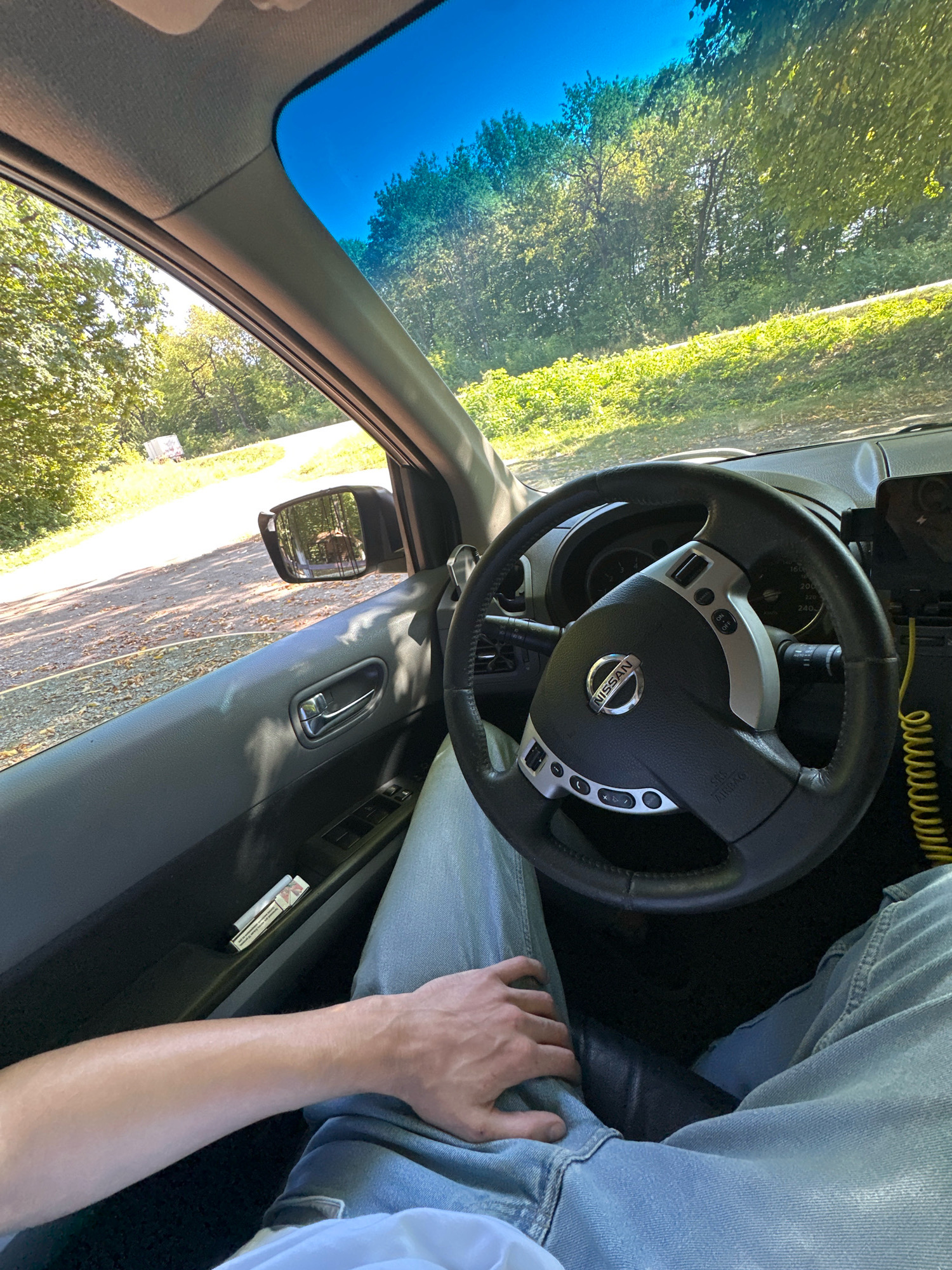 The image depicts the interior of a Nissan car, viewed from the perspective of the driver. The driver's arm and legs, clad in light blue jeans, are visible. A pack of cigarettes rests on the driver's side door panel. The steering wheel, bearing the Nissan logo, is in the foreground. Outside the car, through the windshield and side window, a tree-lined rural landscape is visible, with a dirt road and a distant white truck. The scene appears to be in a natural, possibly wooded, area. The specific location in Shatava, Ukraine, is not discernible from the visual information alone, as no identifying landmarks or signs are present.