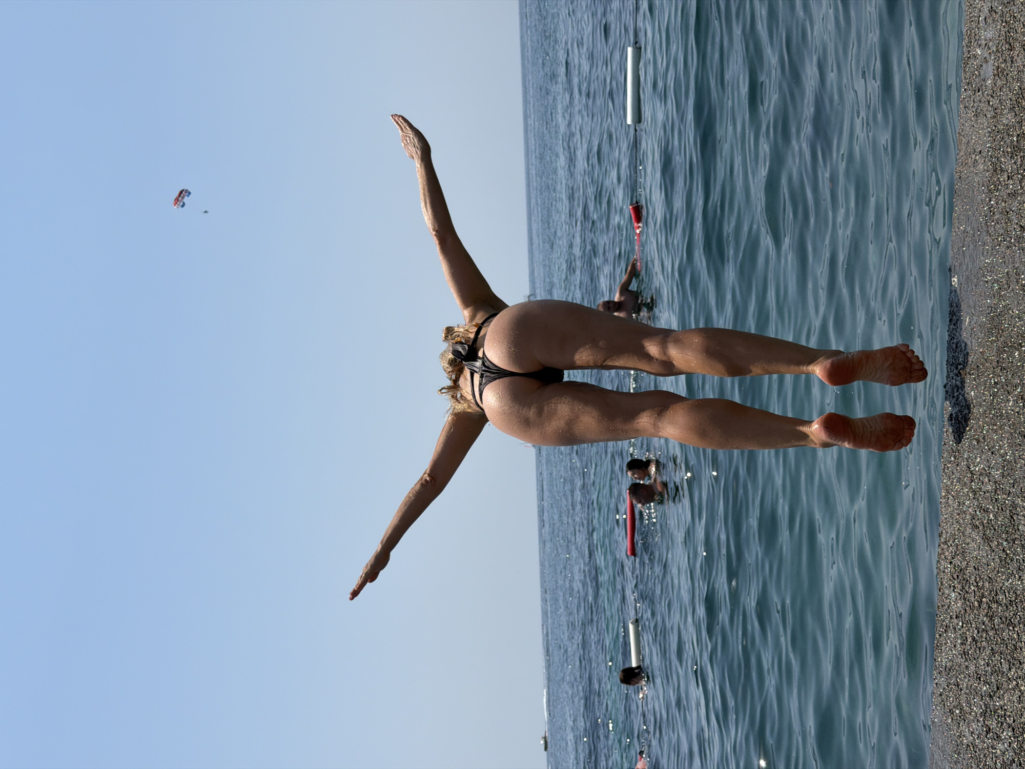 The image captures a woman in mid-air, performing a split leap on a beach in Budva, Montenegro. She is wearing a black bikini, her arms outstretched in opposite directions and her legs spread wide. The setting is a sun-drenched beach with calm, blue waters of the Adriatic Sea. The sea is marked by buoys and ropes, suggesting a designated swimming area. In the background, several people are visible swimming and enjoying the water. Further in the sky, a paraglider is seen against a clear blue sky, hinting at a bright, sunny day. The overall mood is one of exhilaration and freedom, capturing a moment of active recreation on a beautiful summer day. The image is oriented vertically, with the sky on the left and the water and beach on the right.