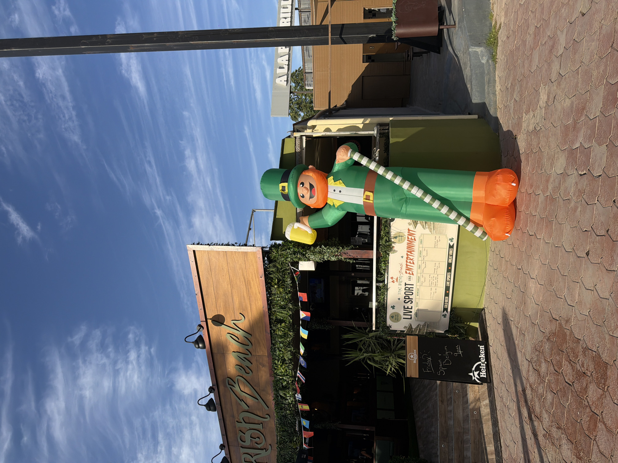 This image shows the exterior of "The Irish Beach" bar in Sant Antoni de Portmany, Spain, on a sunny day with a blue sky and wispy clouds. A large, inflatable leprechaun holding a beer is prominently displayed in front of the establishment. The bar features a wooden facade with the name "Irish Beach" in cursive lettering and is decorated with a string of international flags. To the right of the leprechaun, a sign reads "ALAP ... RILL".
In front of the bar, a blackboard menu lists events, including "Friday?" with "Spain v Belgium" at "9pm", along with the Heineken logo. Another larger sign, also featuring shamrocks, advertises "THE IRISH BEACH LIVE SPORT AND ENTERTAINMENT" with a weekly schedule. The ground in front of the bar is paved with reddish-brown hexagonal tiles, casting long shadows from the objects and architecture. To the right of the scene, a dark metal pole stands tall against the sky. The overall environment suggests a lively entertainment venue.