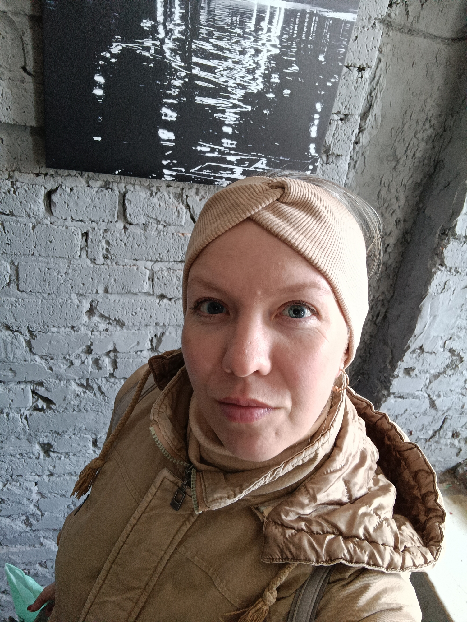 A head-and-shoulders portrait captures a woman indoors, likely in Chaikovsky, Russia. She appears to be in her 30s to 40s, with light skin, blue eyes, and a neutral, calm expression as she looks directly at the camera. Her light-colored hair is partially visible beneath a light beige, ribbed, knotted headband. She is dressed in a matching light beige turtleneck or high collar and a light brown or tan quilted jacket with visible zippers and drawstrings. A small, golden-toned hoop earring is visible on her right earlobe.
The background consists of a textured, white-painted brick wall covering the left side of the frame and behind her. To the right, a darker, rough-hewn concrete or plastered wall/pillar is visible. Hanging on the brick wall behind her left shoulder is a rectangular piece of black and white artwork, featuring abstract or blurred white patterns and lines on a dark background, possibly depicting water reflections or urban lights. A small, light green object, potentially a bag or container, is partially visible in her hand at the bottom left of the frame. The lighting suggests an indoor setting with ambient light. No discernible text is visible.