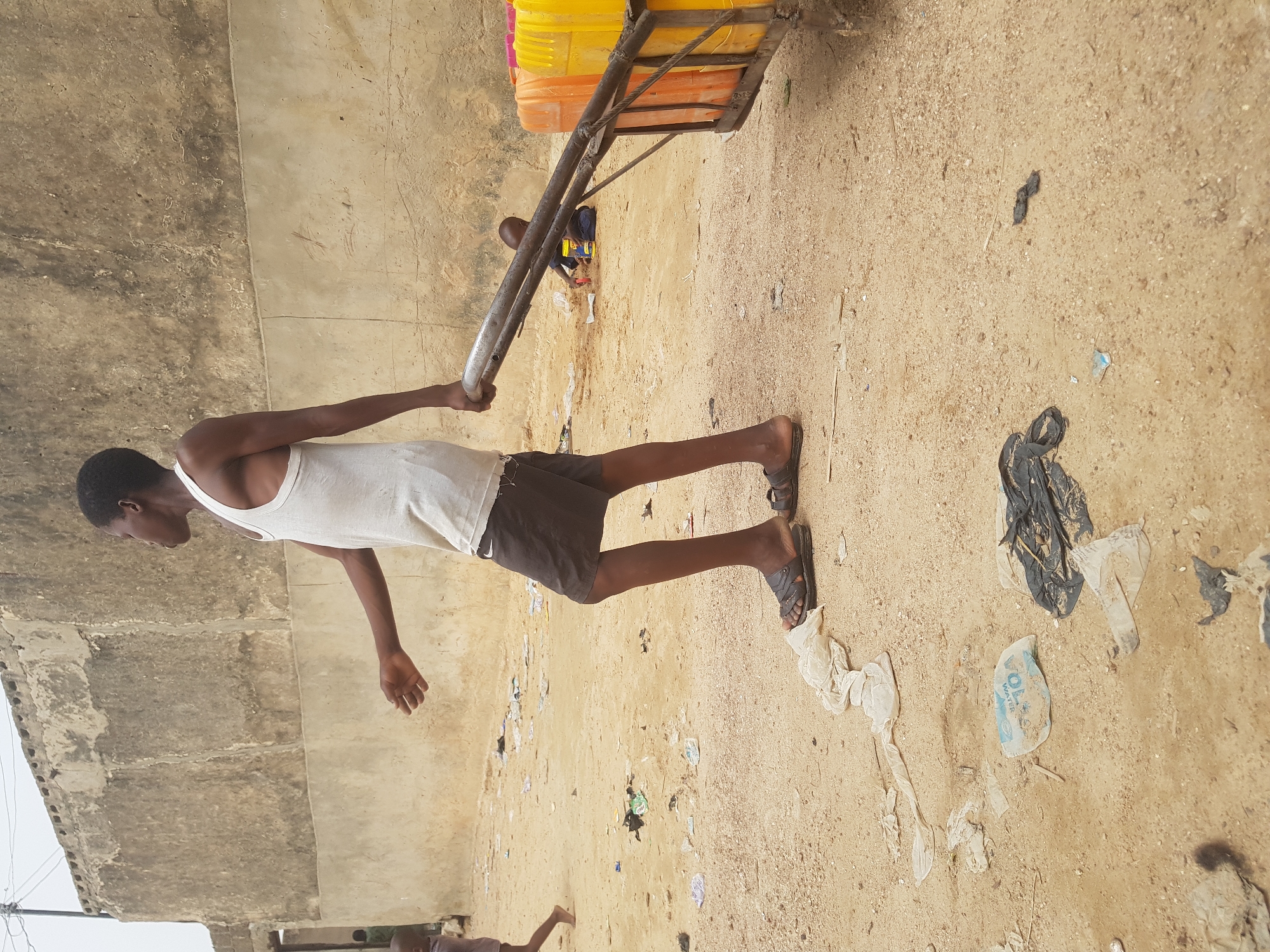 A young man, wearing a white tank top and dark shorts with sandals, pulls a cart laden with stacked yellow and orange containers. A small child, wearing a dark shirt and seated on the cart, appears to be holding a yellow container. The scene is outdoors on a dusty ground littered with trash, including plastic bags. A concrete wall is visible on the left, partially obscuring the sky. The setting appears to be a street in Dutsinma B, Nigeria.
