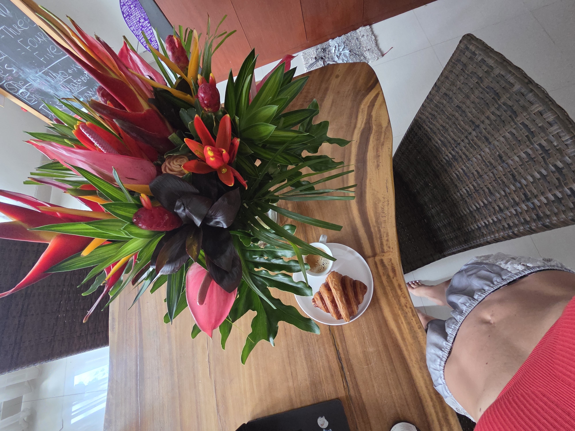 Here is a detailed description of the image:
The image is an overhead shot taken indoors, focusing on a wooden table adorned with a lush, colorful floral arrangement. The arrangement features vibrant red, orange, and pink tropical flowers, including heliconias and anthuriums, mixed with green foliage. Beside the floral display, there is a white plate with a croissant and a small cup of coffee.
The table's wood grain is visible, suggesting it's a natural or stained wood surface. In the background, a portion of a chalkboard with chalked writing is visible, adding a casual, perhaps educational, element to the scene. A wicker chair can be seen next to the table, with a person seated. Part of the person's lower body is visible. They are wearing a red top and light gray silk shorts.
Overall, the image conveys a relaxed, tropical, and possibly domestic setting, perhaps a breakfast or brunch scene.
