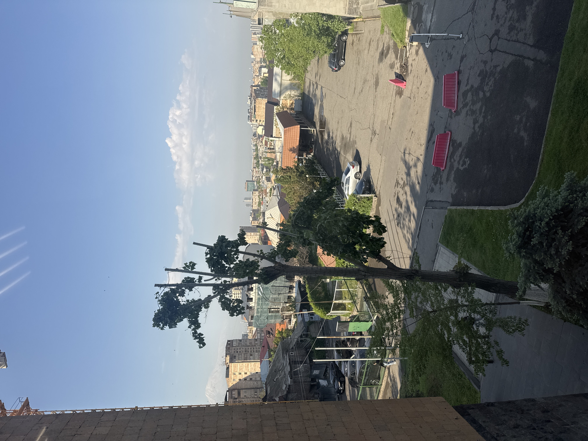 The image is a daytime outdoor shot taken from an elevated position in Yerevan, Armenia. A partly pruned tree is visible in the midground. Beyond the tree, the urban landscape includes buildings, rooftops, and streets. There are cars parked on the cracked asphalt, two red plastic barriers, a red safety cone, and a metal pole with attachments at the top. The sky is blue with a few clouds. The foreground consists of a portion of a brick structure.