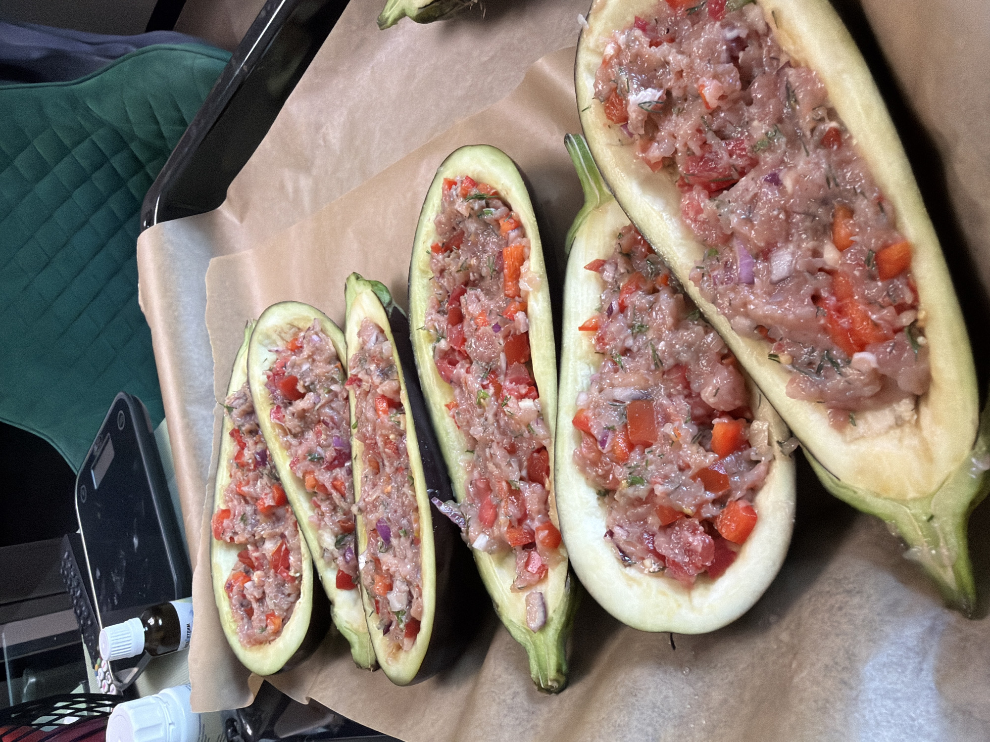 The image shows a close-up, high-angle shot of several halved eggplants stuffed with a mixture of ground meat, diced tomatoes, red onions, and herbs. The eggplants are arranged on a baking sheet lined with parchment paper. In the background, a black digital scale and small bottles are visible on a surface, suggesting a kitchen or food preparation area. The overall lighting is natural and diffused. There are no people or animals present in the image. The scene depicts a food preparation process. This image is not set in Moscow, Russia, and there is no information to suggest it.