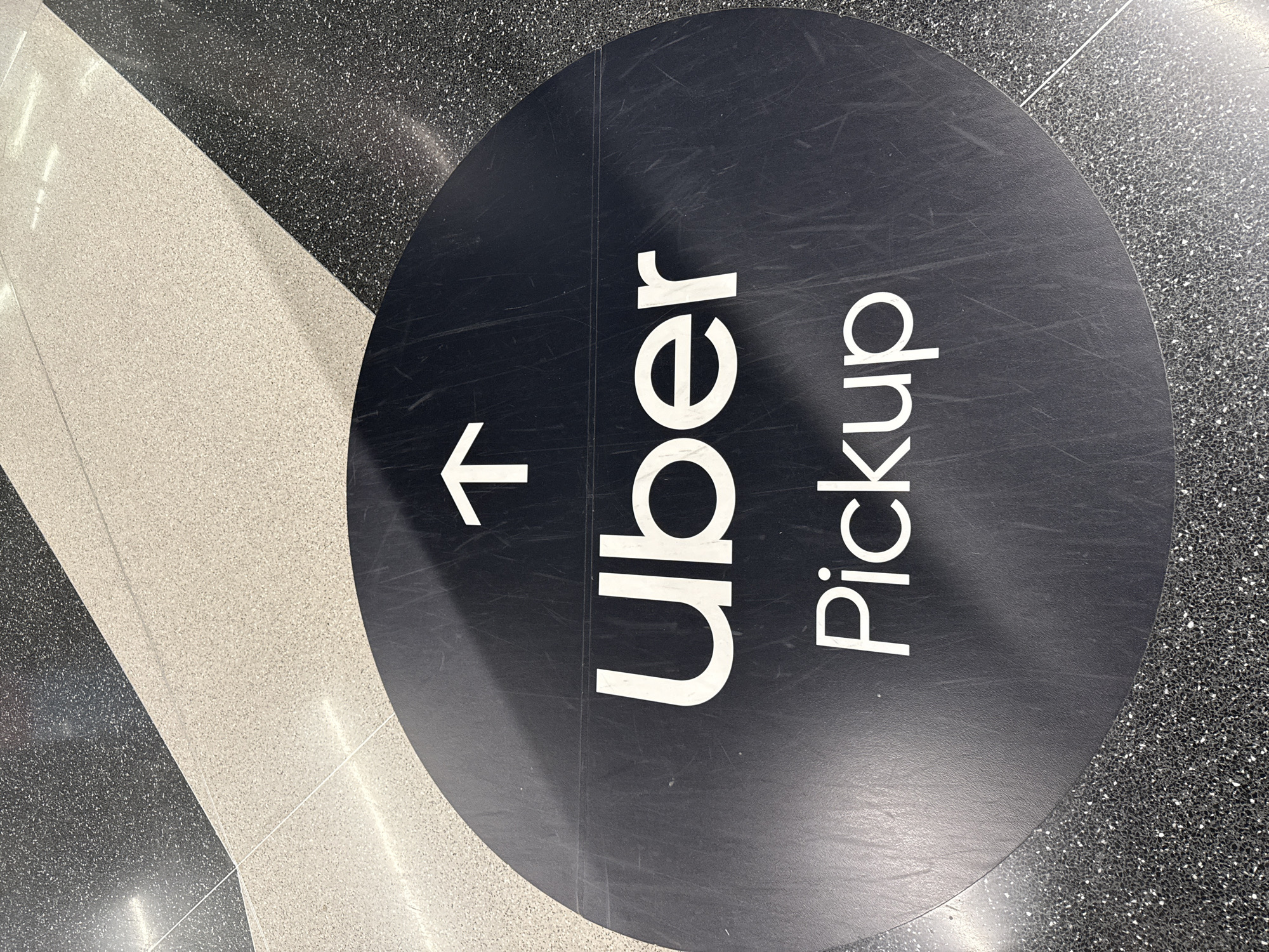 The image depicts an Uber Pickup sign on the floor of what appears to be a transportation hub. The circular sign, primarily black with white lettering, indicates "Uber Pickup" with an arrow pointing to the left. The sign is positioned on a floor with contrasting speckled grey and white segments. There are no people visible in the image. The location is not specified but based on the characteristics of the floor it is an area in the city of Chicago, United States of America.