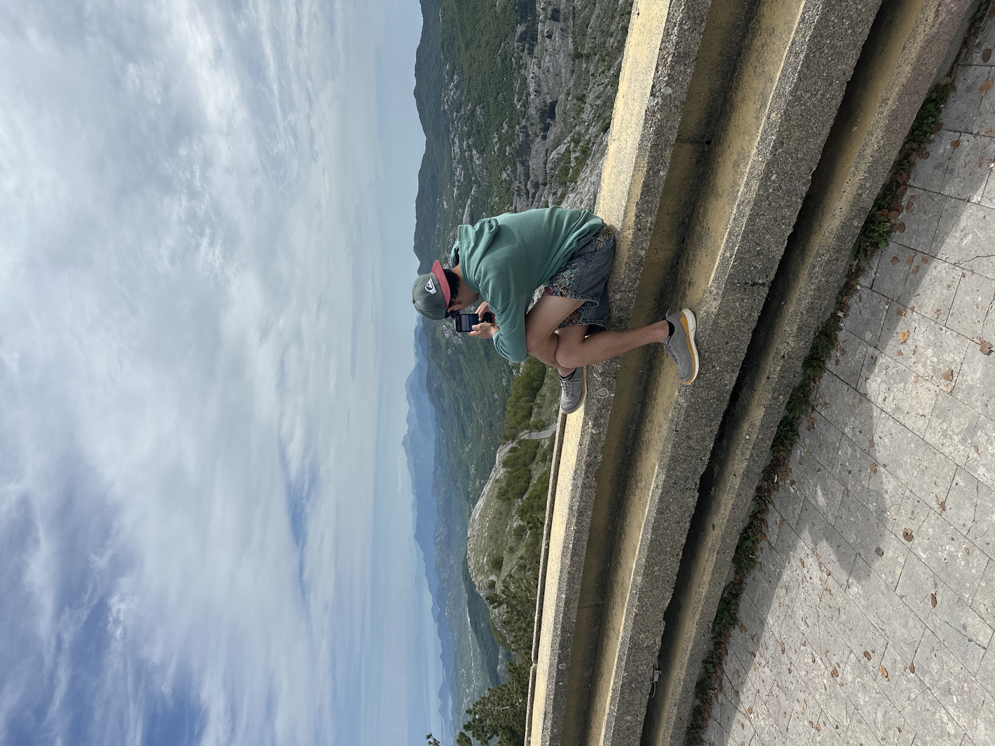 A medium shot captures a person perched on a concrete barrier, holding a mobile phone to take a photograph of the mountainous landscape. The person is wearing a light green hoodie, patterned shorts, and grey shoes. The environment consists of a vast mountain range under a cloudy sky. The concrete barrier is a structure with multiple horizontal bars, providing a safety measure at the edge of what appears to be a scenic overlook. The location is identifiable as Mali Bostur, Montenegro, due to the characteristic mountainous terrain visible in the background. The action of the person indicates that they are documenting or capturing the scenery with their phone.