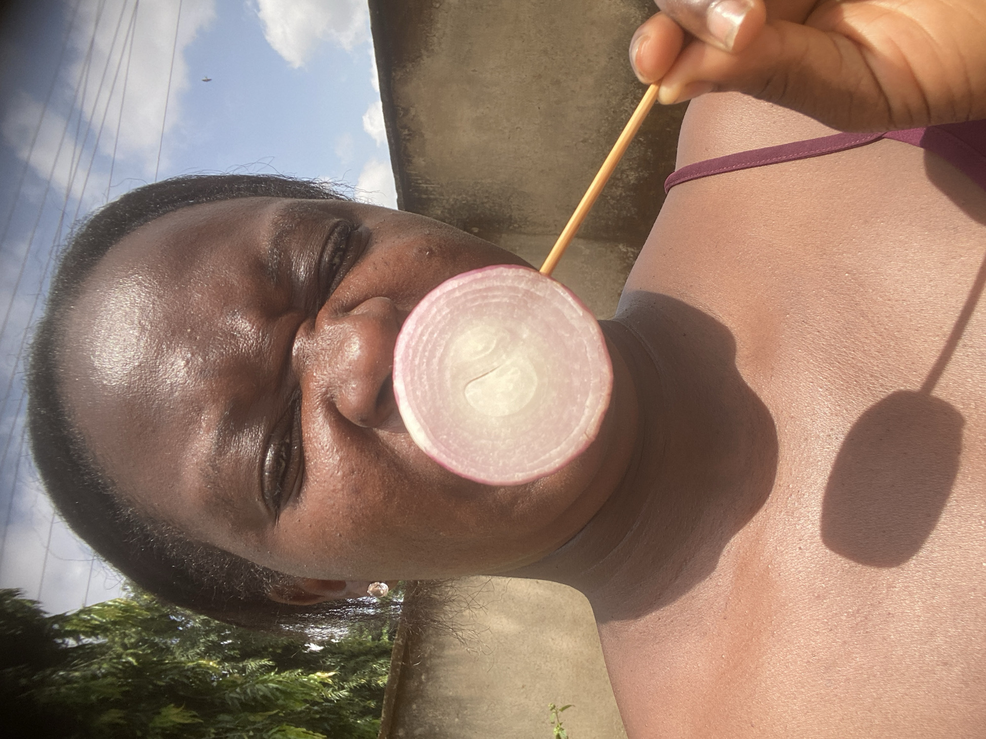 This close-up, portrait-style image captures a person outdoors in Maigizo, Nigeria, under bright, sunny conditions. The person, with dark skin and short dark hair, is positioned such that their head and upper torso fill a significant portion of the frame. They are holding a thin, circular slice of red onion on a wooden skewer directly in front of their mouth and nose area. The onion slice displays its distinct concentric layers and a light pink hue.
The person's right hand, with light-colored fingernails, is visible in the upper right, gripping the skewer. A maroon or dark red strap, likely from clothing or swimwear, is visible on their right shoulder and upper chest. The strong overhead sunlight casts sharp shadows, including a noticeable shadow from the onion and skewer on the person's chest.
In the background, a clear blue sky with sparse white clouds is visible, suggesting good weather. A light-colored concrete or stucco wall or structure forms a substantial part of the background, particularly behind the person's head and shoulder. Green foliage, possibly trees or bushes, can be seen in the lower-left corner, and faint power lines are visible against the sky in the upper left. The person's expression, partially obscured and squinted, hints at a playful or lighthearted moment. There is no visible text in the image.