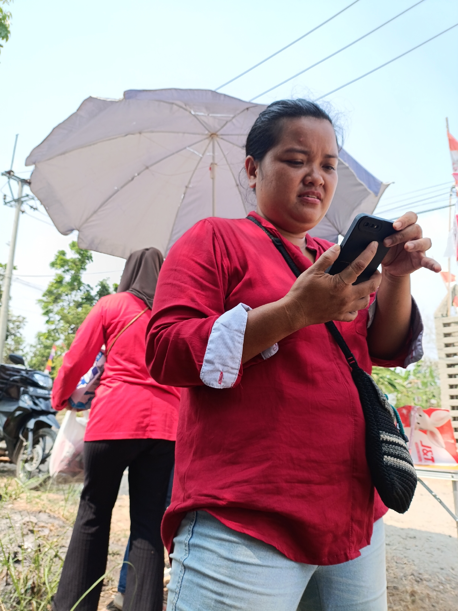 A woman in a red shirt and light blue jeans is standing outdoors, holding a black smartphone and looking at its screen. She has dark hair pulled back and is wearing a dark strap bag over her shoulder. Behind her, another person in a red shirt and dark pants is visible, carrying a white bag and wearing a brown headscarf. A large white umbrella is held above them. A motorcycle is parked to the left. The ground appears to be dirt with some dry grass. In the background, there are trees, a utility pole, power lines, and a building with white siding. Red and white flags are also visible. The scene is likely in Cariu, Indonesia, given the presence of local flags and typical outdoor market elements.