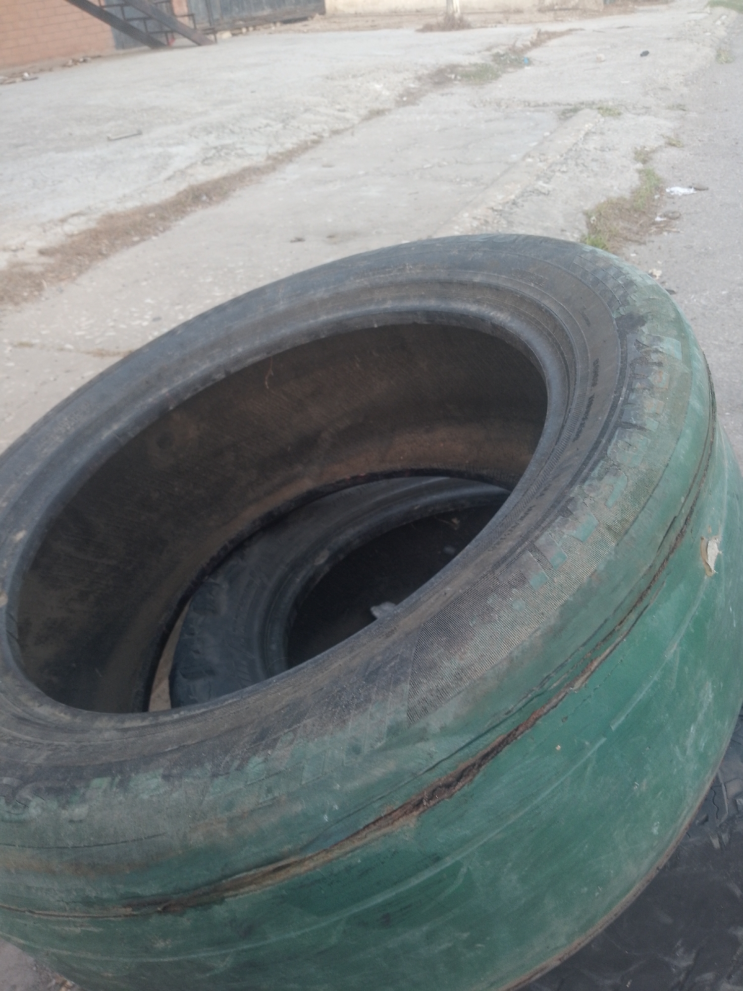 The image shows a stack of old, worn-out tires. The top tire is painted green and has a noticeable crack running along its side. The tires are located outdoors, likely on a paved surface in an urban environment. The background shows a concrete road and a partially visible metal structure, potentially a gate or scaffolding. The exact location in Porlamar, Venezuela, cannot be determined from the image alone.