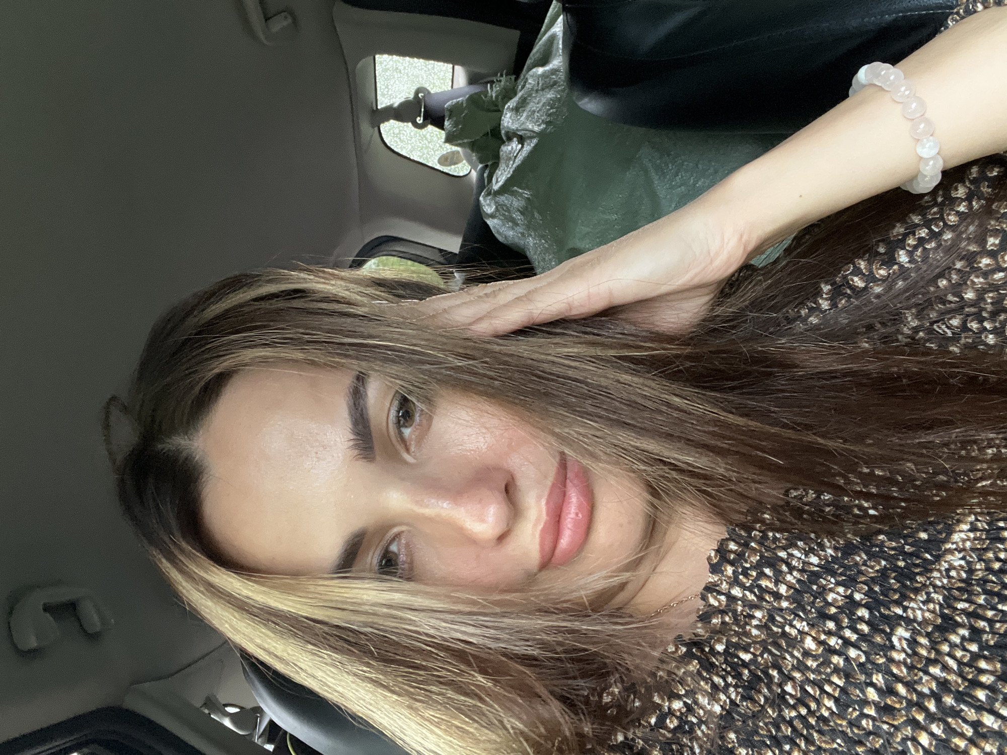 A young woman with long, layered brown and blonde hair and brown eyes is in the passenger seat of a car. She is wearing a dark top with a small, abstract pattern. She is resting her right hand on her head and looking at the camera. A green tarp is visible in the back seat, covered with items. The weather outside appears to be rainy.