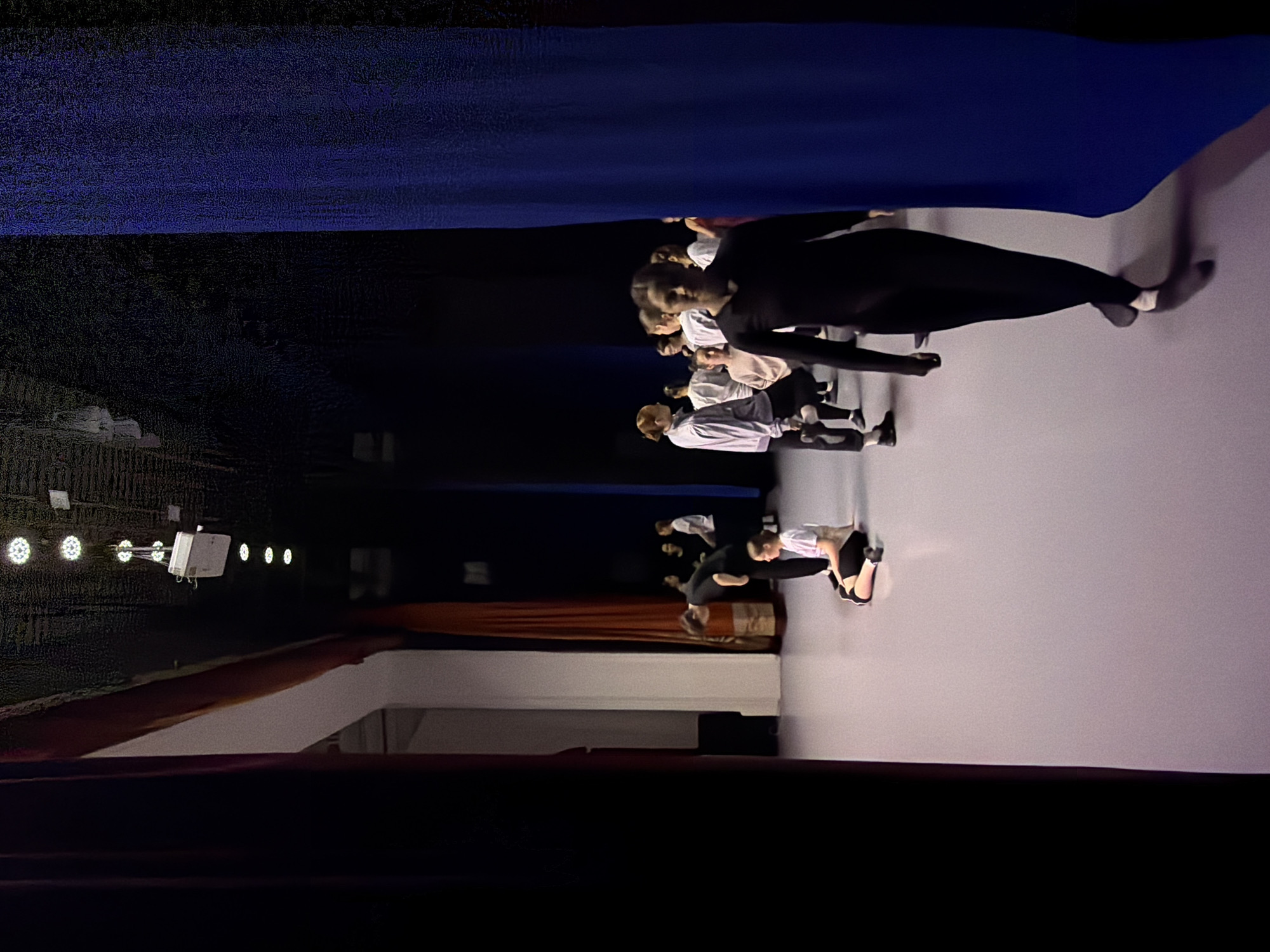 This media file captures an indoor scene, likely a dance studio or performance space, in Perm, Russia. The image appears to be rotated 90 degrees clockwise, so we will describe it as if rotated counter-clockwise to an upright position.
The setting features a large blue fabric or curtain spanning the top of the frame, possibly as a backdrop or ceiling drape. Below this, a dark, textured wall serves as the background. On the lower portion of the image, there is a smooth, light-colored floor, likely white or light grey, on which the subjects are positioned.
A group of approximately eight to ten young individuals, presumably dancers or performers, are visible on this light floor. They are dressed in activewear, predominantly black leggings or tights, often paired with white or light-colored tops and some in all-black ensembles. Their postures suggest they are engaged in a choreographed activity, such as dancing, posing, or rehearsing. One individual in all-black activewear is prominent in the mid-ground, bending at the waist. Another, closer to the viewer, is seen from behind, also in a dance-like pose.
In the background, on the left side of the dark wall, several circular stage lights are visible, emitting bright, focused illumination. A white rectangular electrical device or fixture is also mounted near these lights. Further to the right, behind the group of people, sections of brown and darker, possibly maroon, fabric or curtains are hanging. Shadows from the performers are cast on the light floor, indicating the direction of the stage lighting.
The overall impression is a focused rehearsal or performance in a controlled, dimly lit environment, characteristic of a dance or theater setting. There is no visible text in the image.