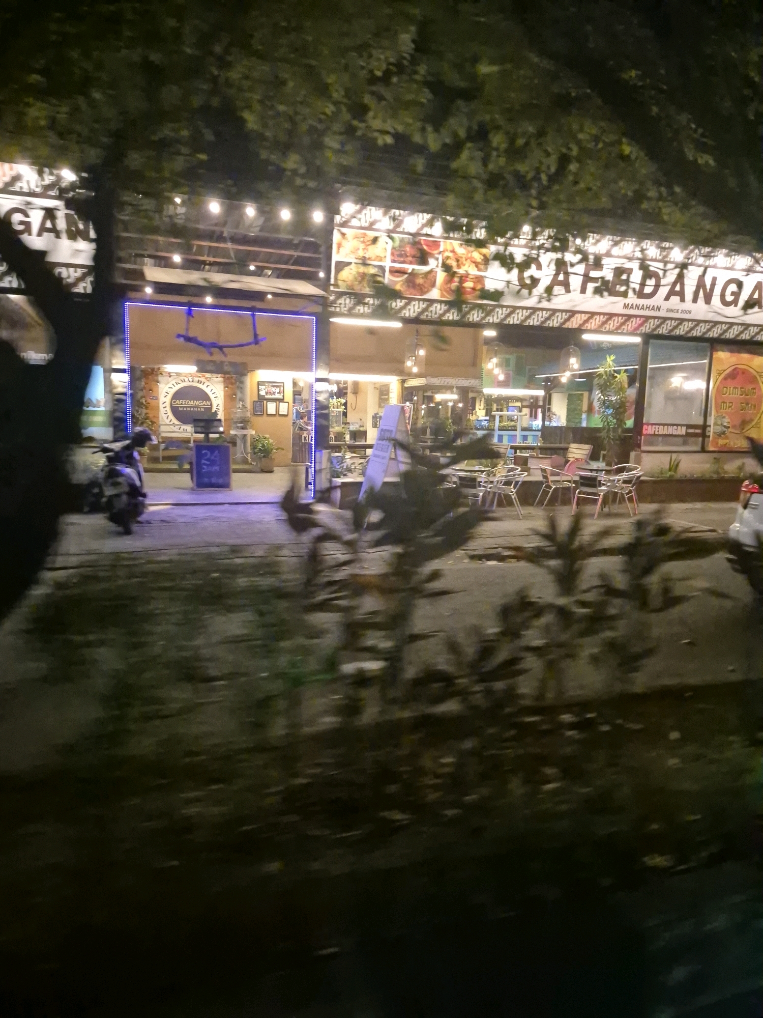 The image shows the exterior of a cafe at night in the Manahan area of Surakarta, Indonesia. The cafe, named "CAFEDANGAN" and also displaying the name "GANDA" on a sign, is illuminated by string lights and the neon blue glow of its entrance. Outdoor seating with metal chairs and tables is visible in front of the cafe. A motorcycle is parked to the left of the entrance. The cafe appears to be busy with patrons visible inside. The overall scene depicts a lively nighttime atmosphere in a commercial district.