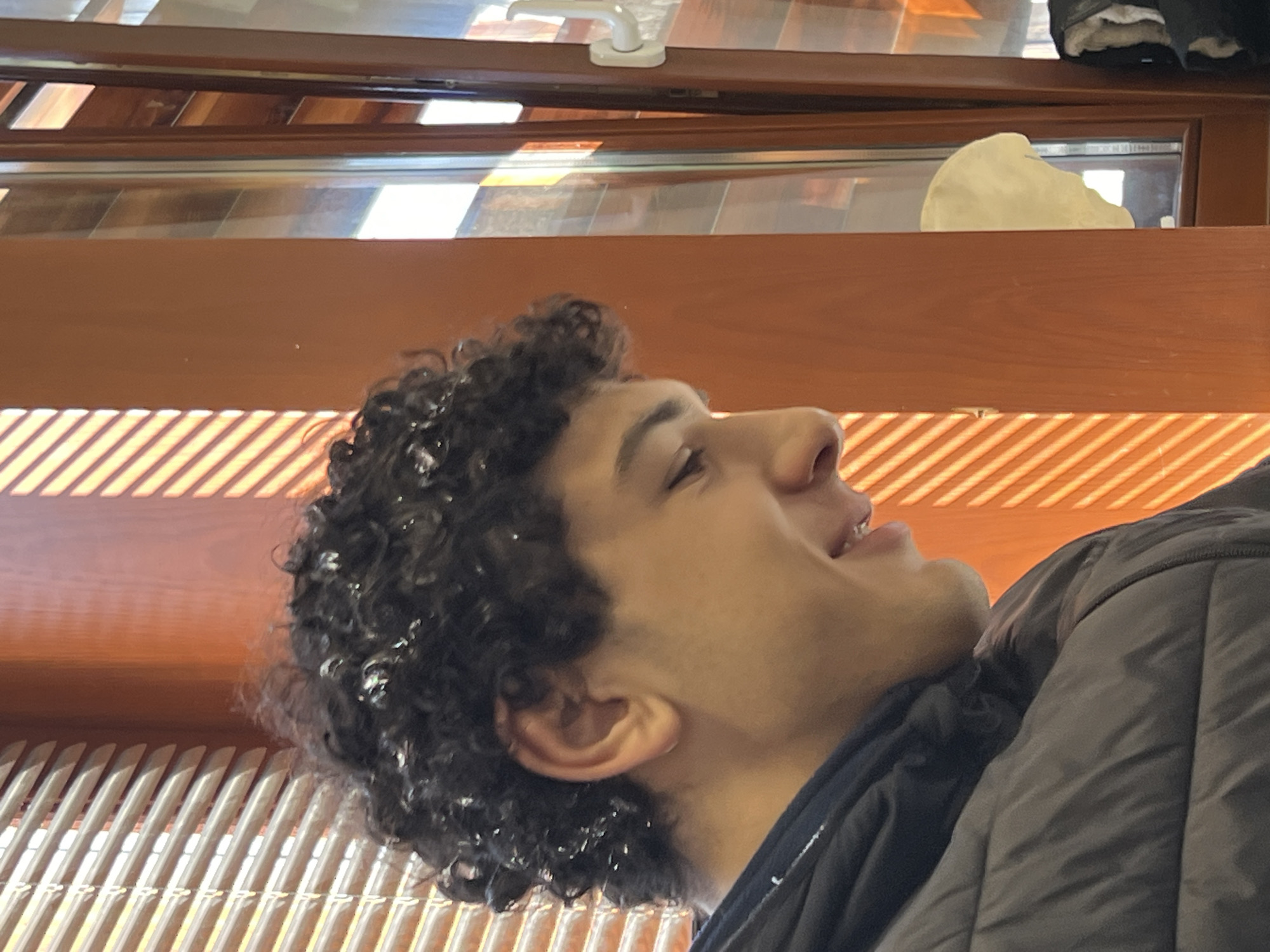 A close-up, low-angle shot shows a young man with dark curly hair, facing right and looking upwards. He is wearing a dark jacket. The background consists of a wooden window frame and what appears to be the slats of blinds or shutters with sunlit stripes across them. Above the window frame are glimpses of the exterior. The location is suggested to be Schiltigheim, France.