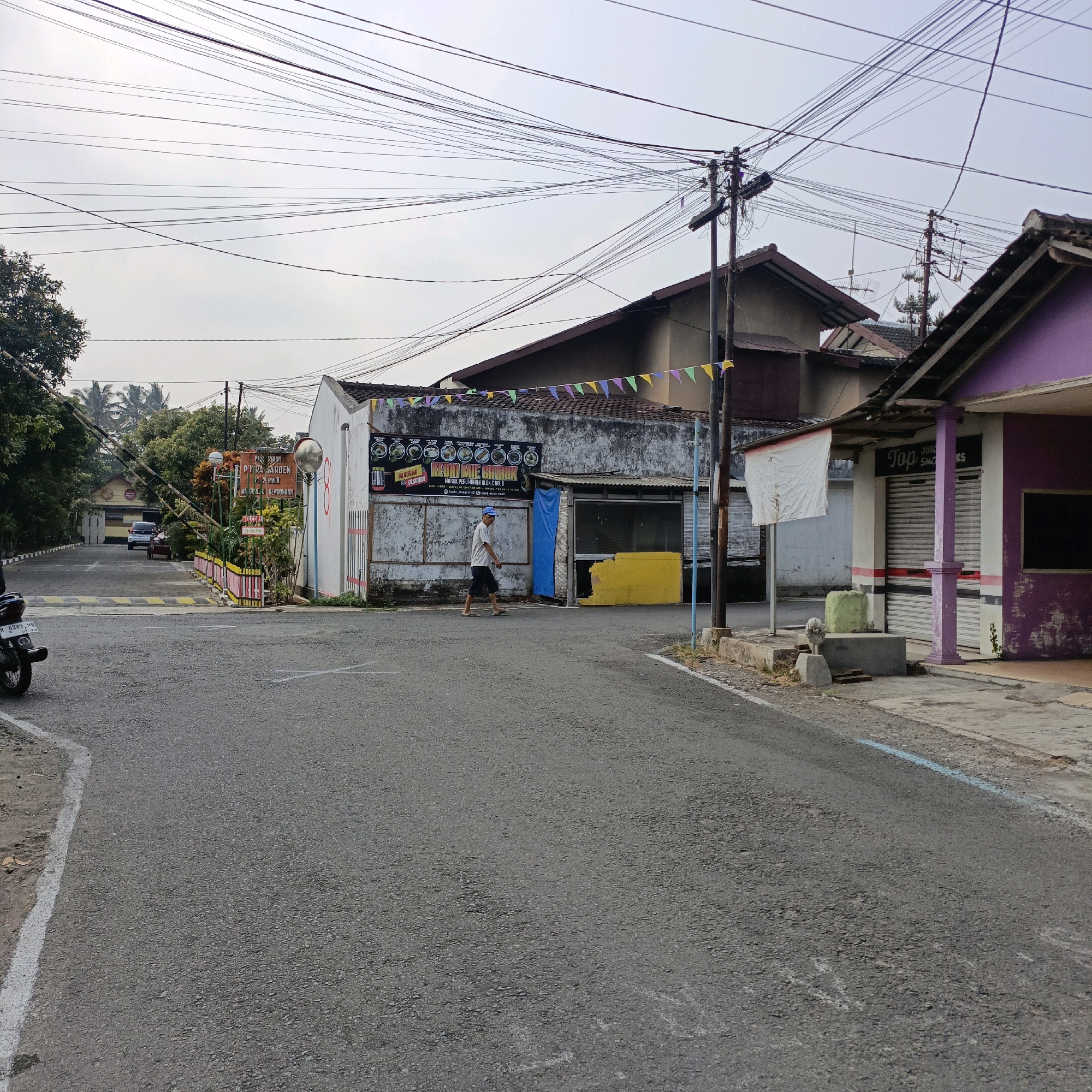 The image depicts a street scene in Salatiga, Indonesia, featuring a mix of commercial buildings and residential structures, overlaid with a dense network of overhead electrical and telecommunication wires. On the right side of the road, a shop with a purple facade and metal roller shutters is visible, with a sign reading "Top JUICE SMOOTHIES". Adjacent to it, another building with a white exterior and similar roller shutters shows a white banner.
In the center of the frame, a man wearing a light-colored t-shirt and dark shorts walks across the road. Behind him is a building with a weathered facade, featuring a large banner advertising "KEDAI KITE CHORUK". The building has a blue tarp covering an opening and a bright yellow structure. Festive triangular flags are strung across the front of this building and the purple-fronted shop.
To the left, a road leads to what appears to be an entrance with a barrier, signage indicating "PT. INDRA GARDEN", and a reflective mirror for traffic. A white car and a small trailer are visible beyond the barrier. A motorcycle is parked on the far left of the image. Trees and other greenery are present in the background. The road surface is asphalt with visible white road markings. The sky is overcast.