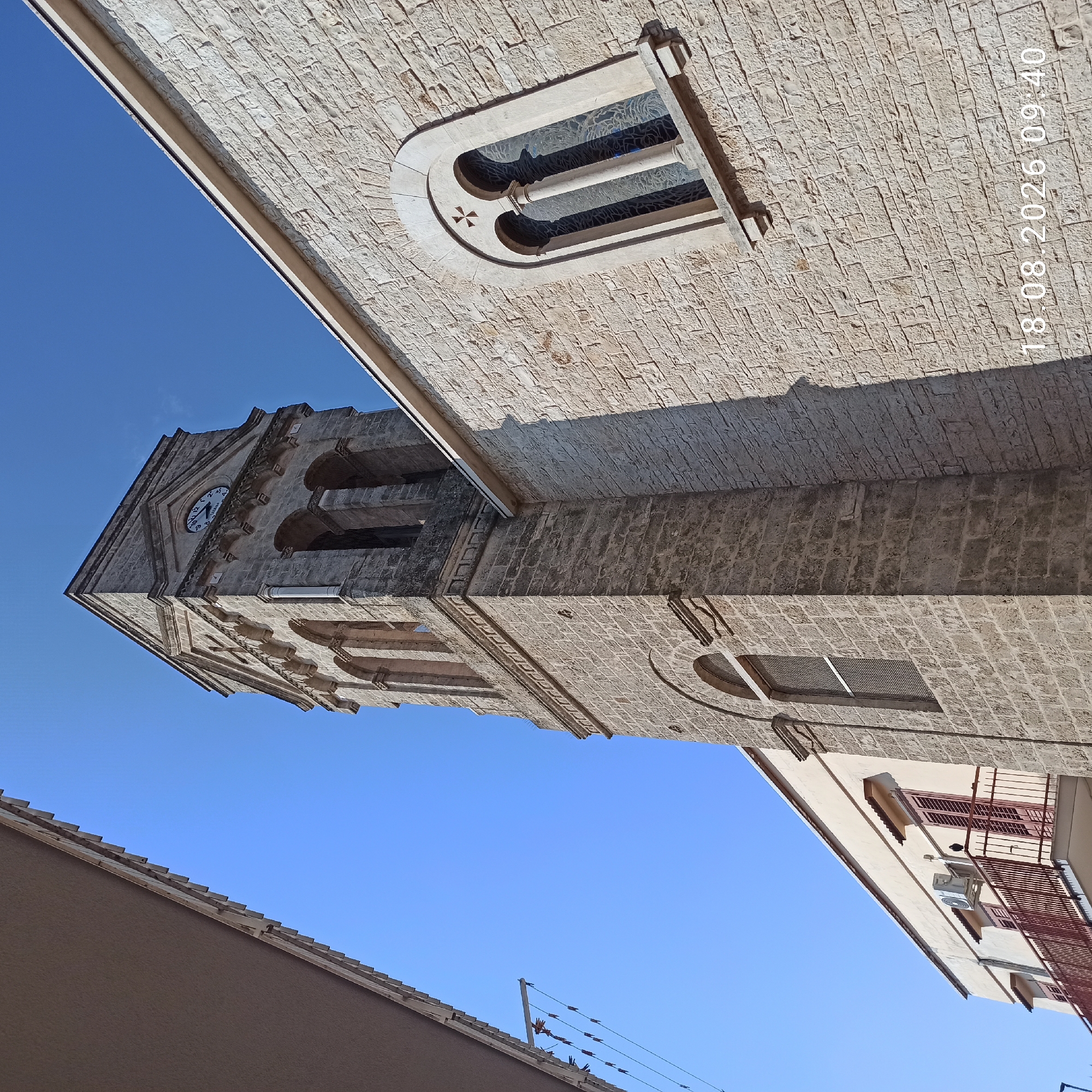 The image shows a low-angle view of a stone church bell tower and part of the adjacent building. The bell tower is constructed of light-colored stone blocks and features multiple arched openings and a clock face. The building to the right is also made of similar stone and has a large arched window with a column dividing it. The sky is clear and blue. In the lower right corner, a timestamp reads "18.08.2026 09:40". A portion of another building, with a balcony and shutters, is visible to the left. The image appears to be taken in the city of Bitonto, Italy, possibly near the Piazza Cavour given the architectural style and the presence of a prominent bell tower. There are no people or animals visible in the image.