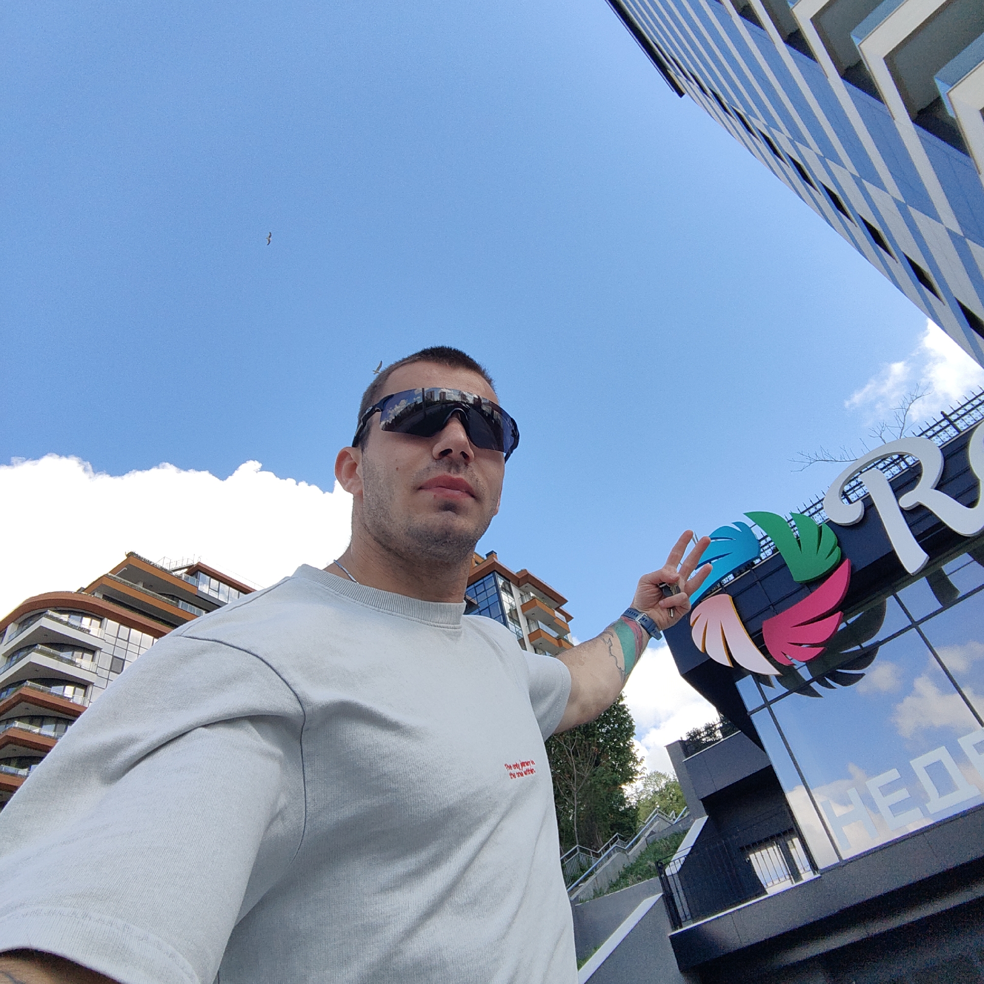 A low-angle shot captures a man outdoors, likely in a city. He is light-skinned with short, dark hair and is wearing sunglasses and a light-colored t-shirt. He is holding up two fingers in a peace sign. Part of a sign with colorful feathers, likely belonging to a business or organization, is next to him. Behind the man are modern buildings and a mostly clear blue sky with a few clouds. A small bird is visible in the sky. The city is Unknown, Unknown.